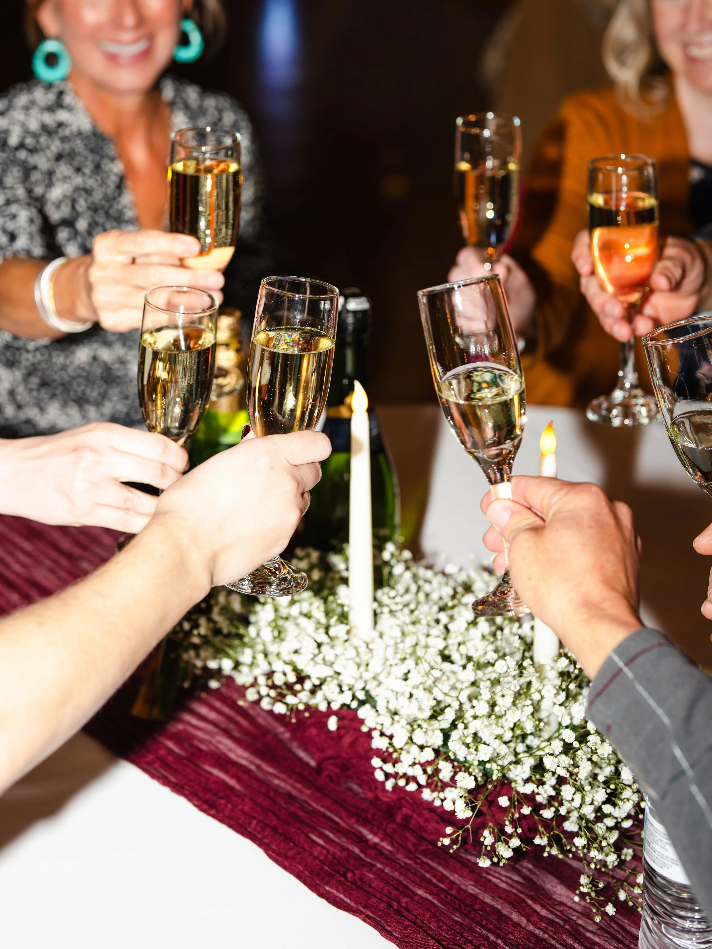 Two women in black graduation gowns and caps exchanging a handshake at a graduation ceremony. One is smiling brightly, holding a red diploma cover, while the other is slightly blurred in the foreground. Other graduates and spectators are visible in the background.