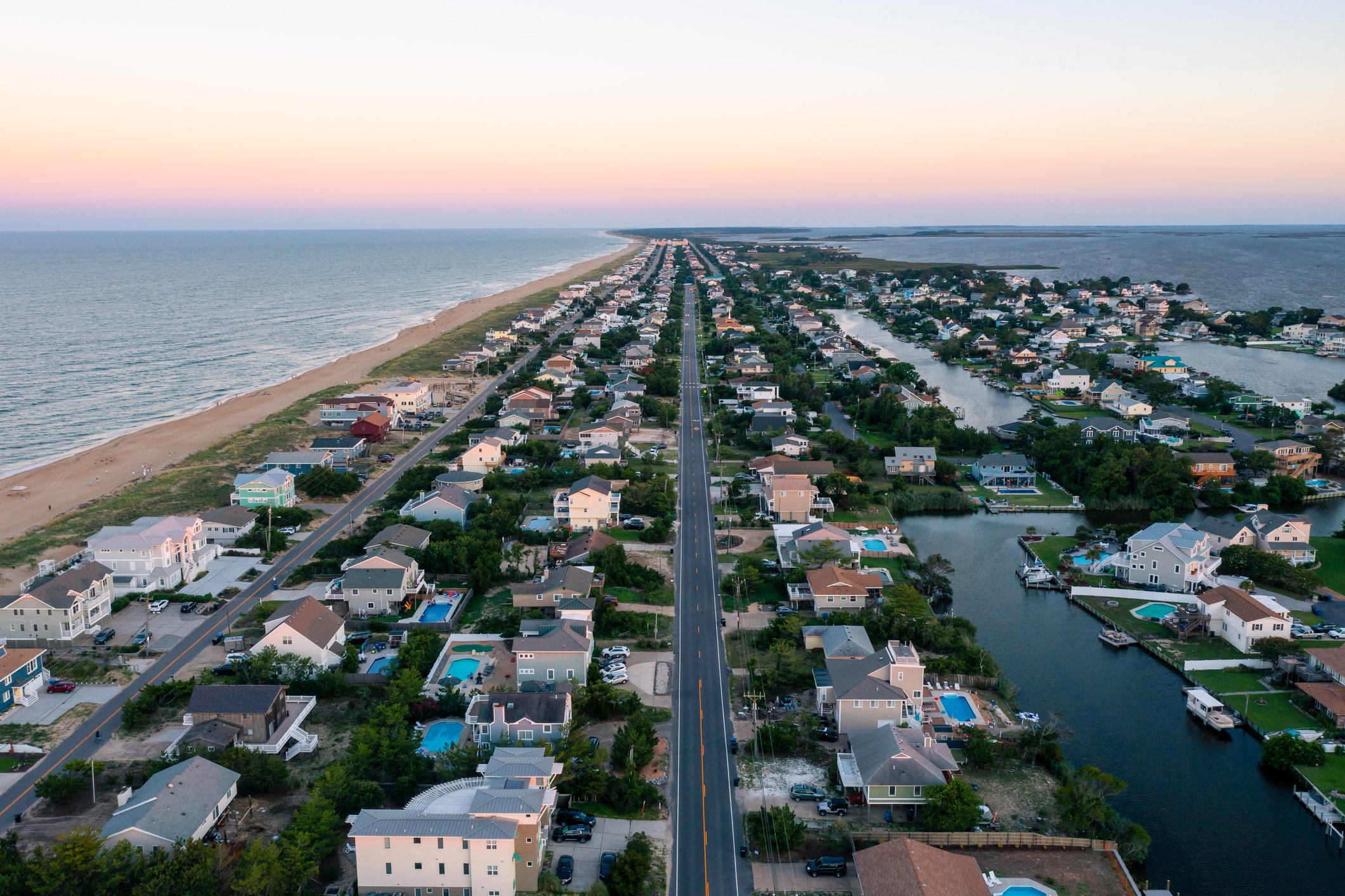 Aerial view of a coastal neighborhood at sunset, showing houses along a canal and a sandy beach on the left, with the ocean extending to the horizon.