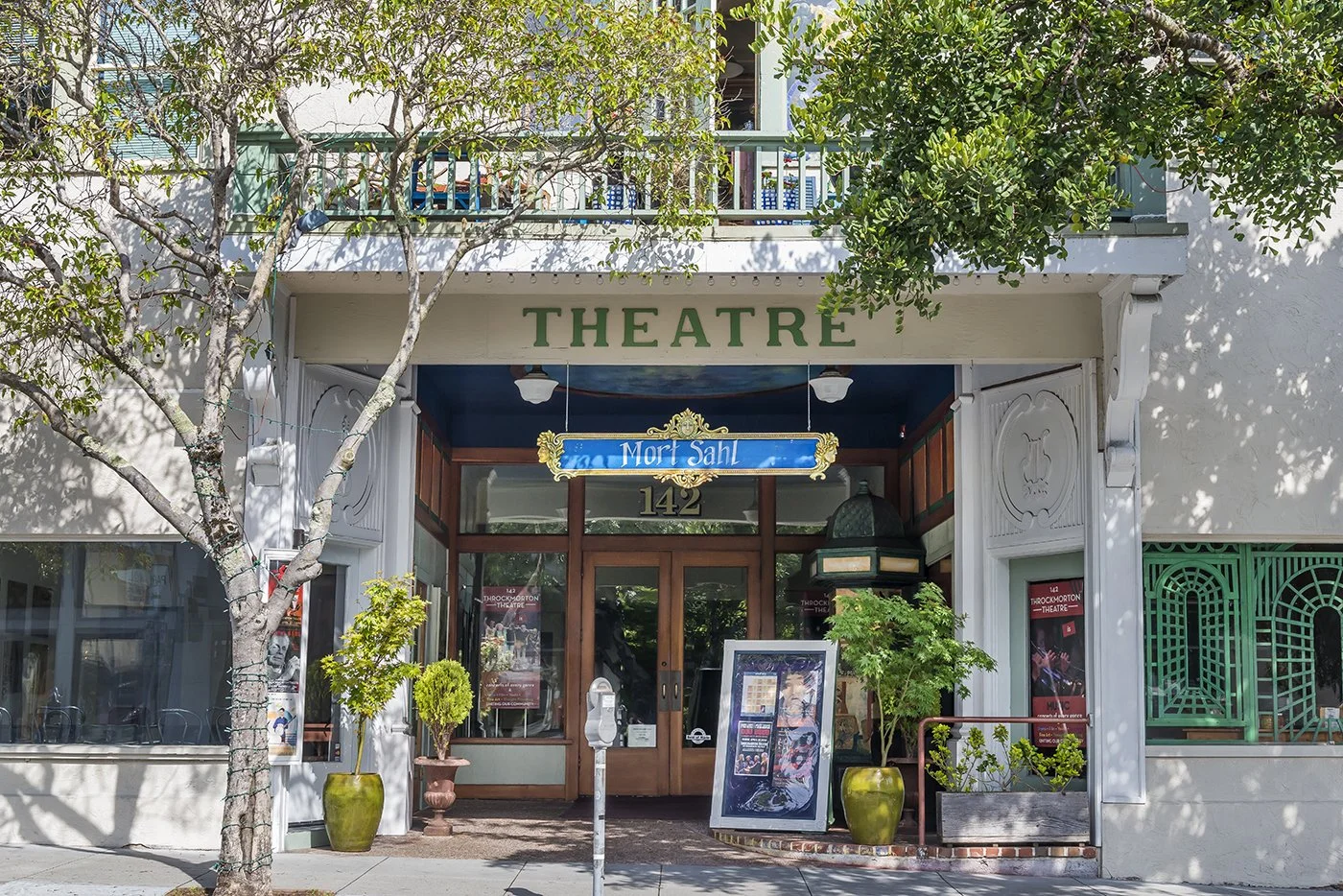 Theatre entrance with green letters, potted plants, a poster display, and a blue sign reading 'Mor Sah' at 142 Throckmorton Theatre.