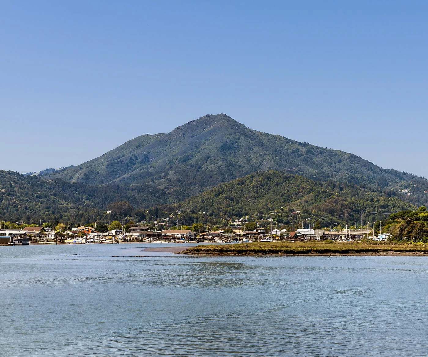 Scenic view of a mountain with lush green slopes, a small town at the base, and water in the foreground under a clear blue sky.