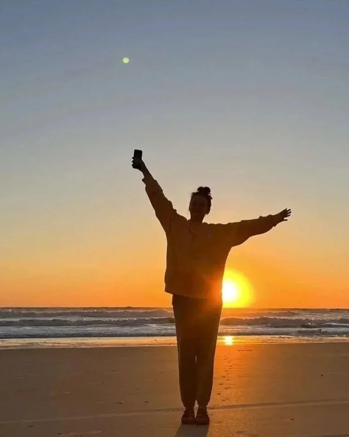 Person standing on the beach at sunset with arms outstretched, taking a selfie with a mobile phone.