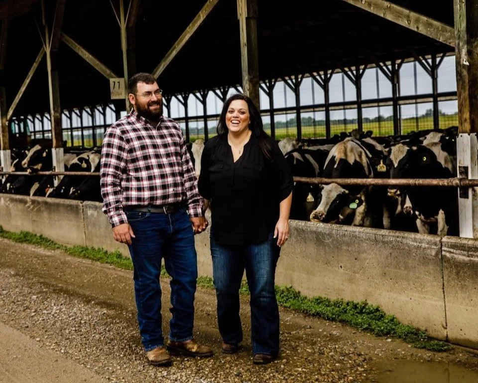 A man and a woman standing hand in hand in a barn or dairy farm, smiling at each other, with cows behind a railing in the background.