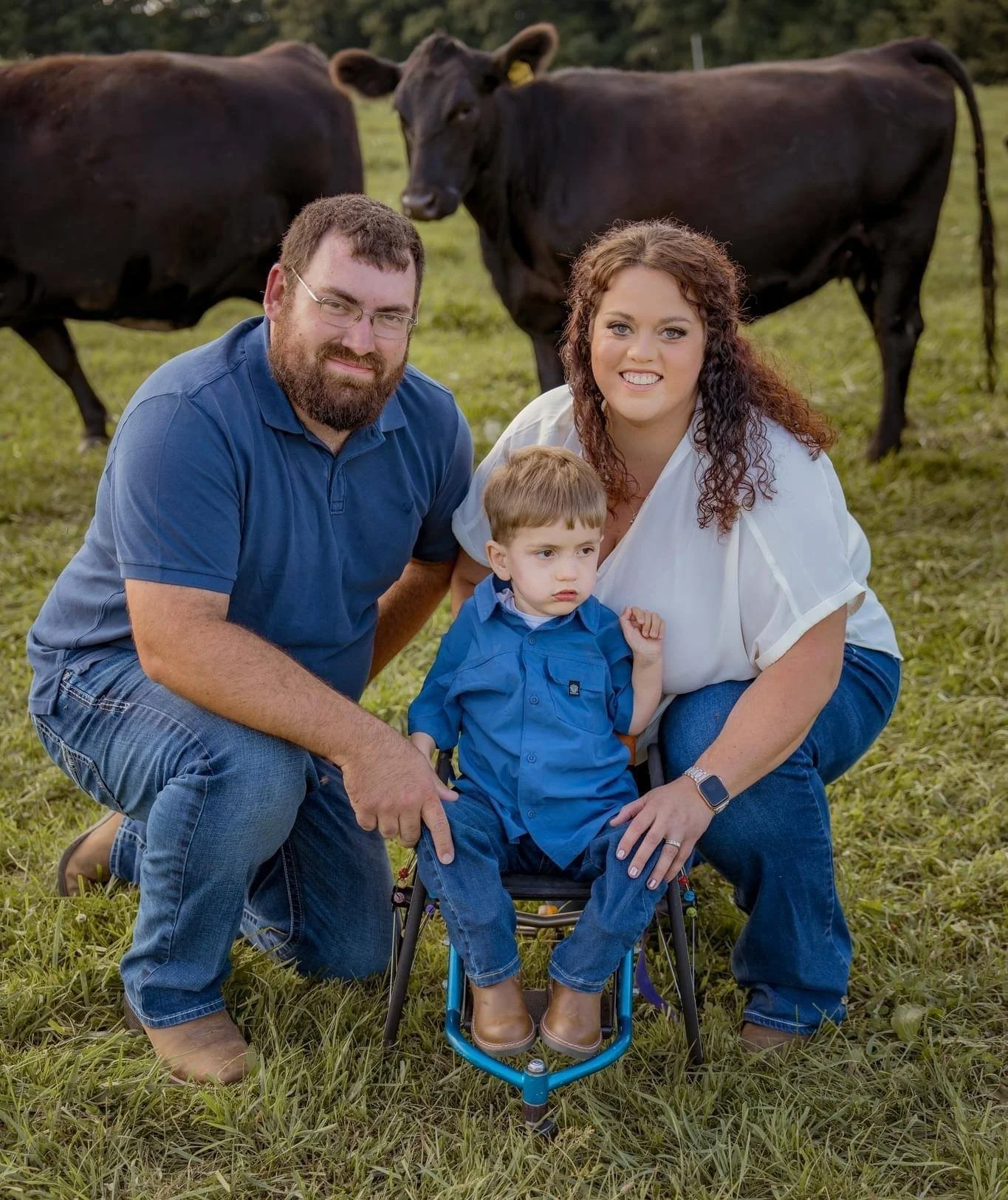 A family of three kneeling on the grass with cows in the background, smiling at the camera.