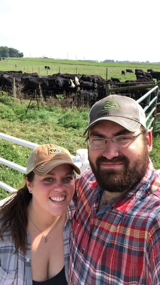 A smiling couple taking a selfie at a farm, with cows grazing in the background and green fields under a bright sky.