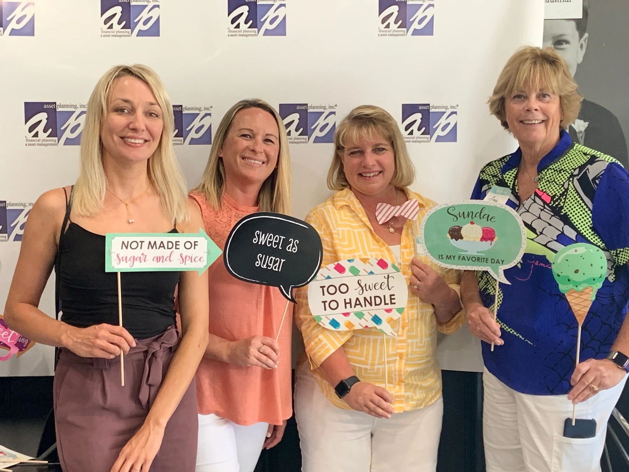 Four women standing together at an event, holding colorful speech bubble signs with messages related to sweets and desserts. The background features a white wall with a logo pattern.