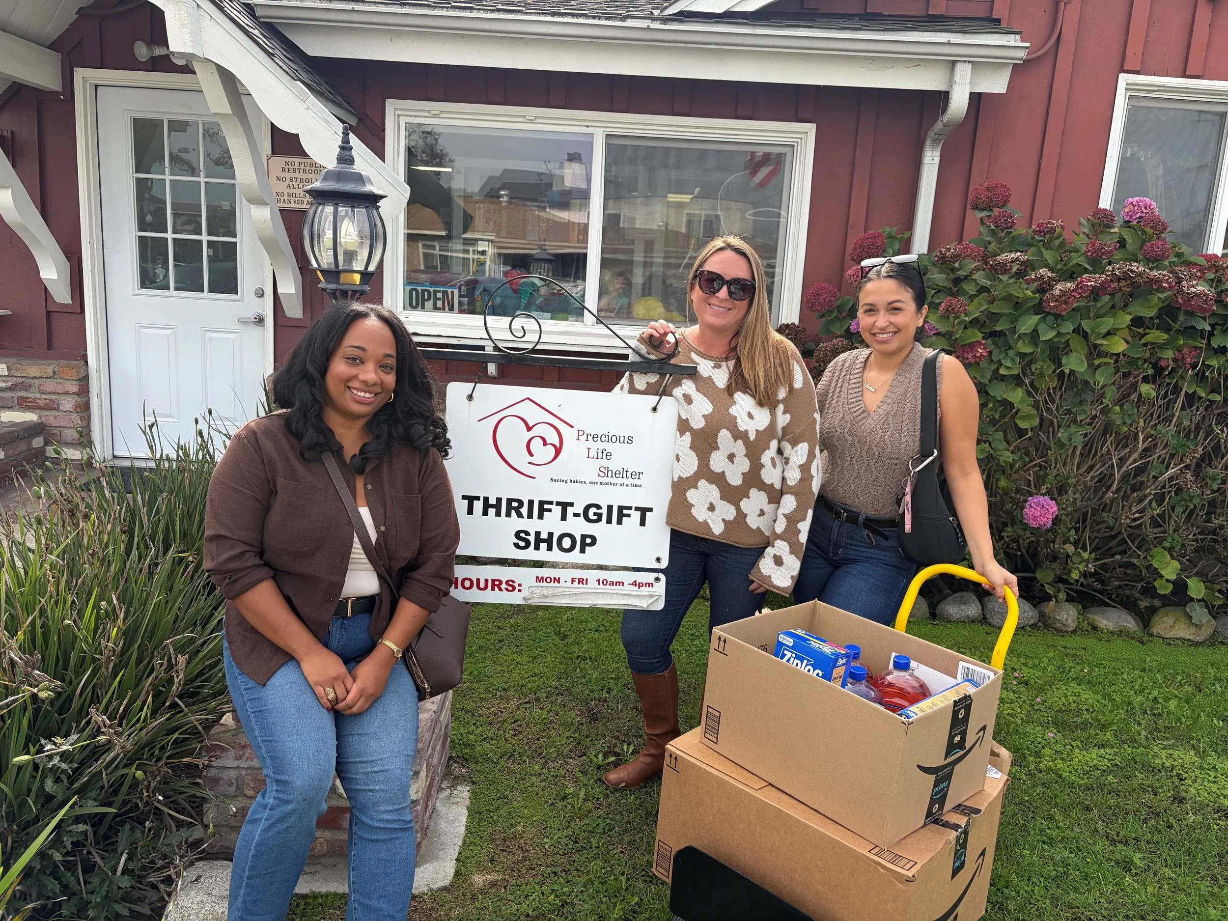 Three women standing outside  Precious Life Shelter, smiling. One woman holds a cart with large boxes of, and they are near bushes with pink flowers. The shop has a red exterior with white trim, a lantern, and an 'Open' sign visible through the window.