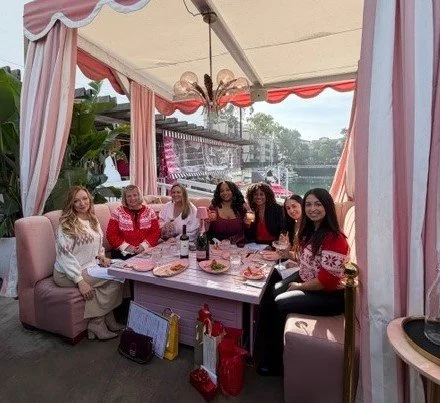 Seven women sitting around a dining table with food and drinks, under a decorated outdoor tent.