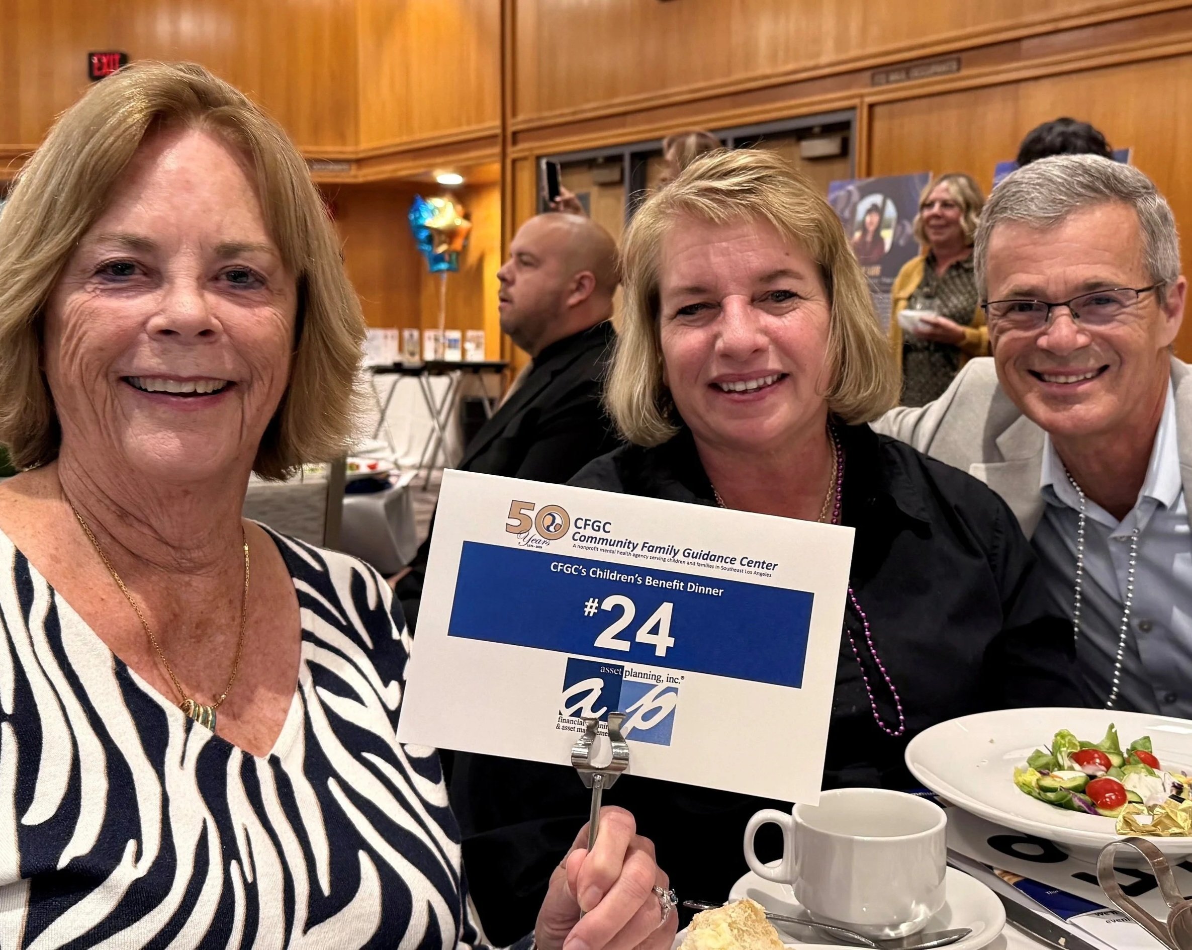 Three smiling people at a dinner event, woman in foreground holding a sign with event details, food on table, wooden-paneled walls in background.