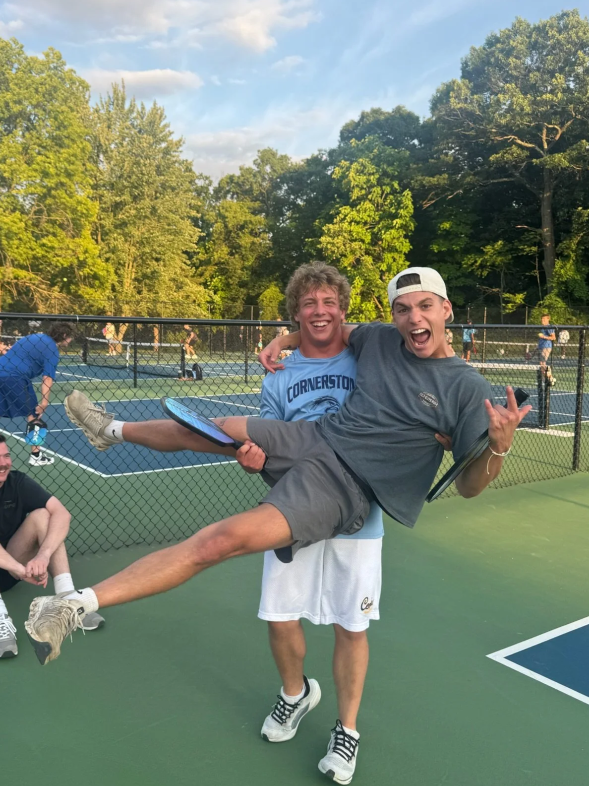 Two young men celebrating on a tennis court, one is holding the other in his arms, both smiling and looking excited, with other people and tennis courts in the background.