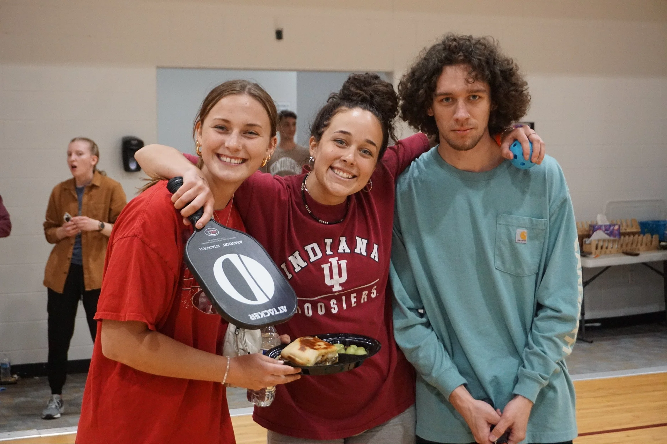 Three young people smiling and posing for a photo indoors. The woman on the left is holding a plate of food and a paddle with a pickleball logo. The person in the middle is wearing a red Indiana University shirt, and the person on the right has curly hair and is wearing a light blue long sleeve shirt. They are standing close together with their arms around each other.