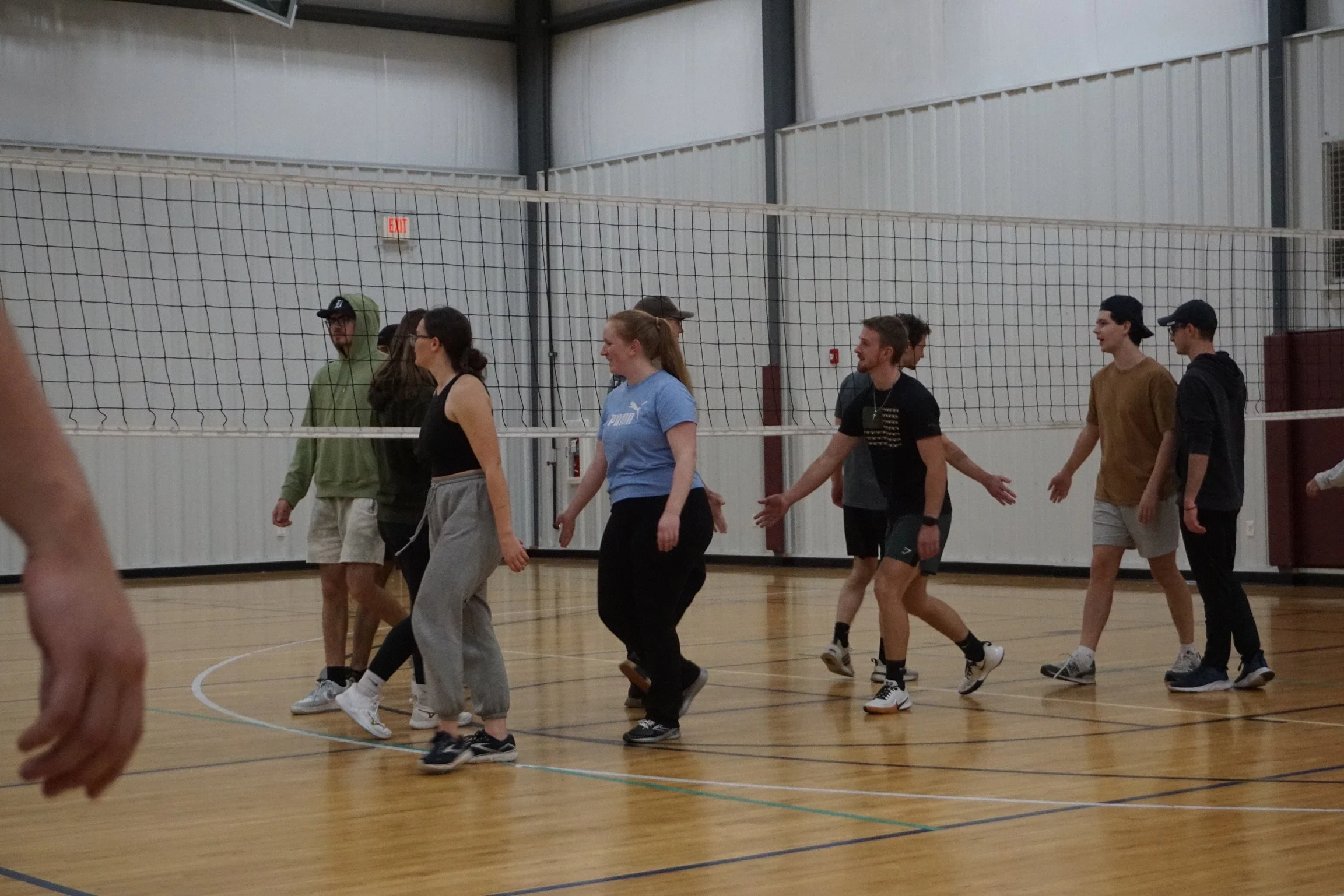 Group of young adults walking on indoor volleyball court, connected by a volleyball net, in a gymnasium.