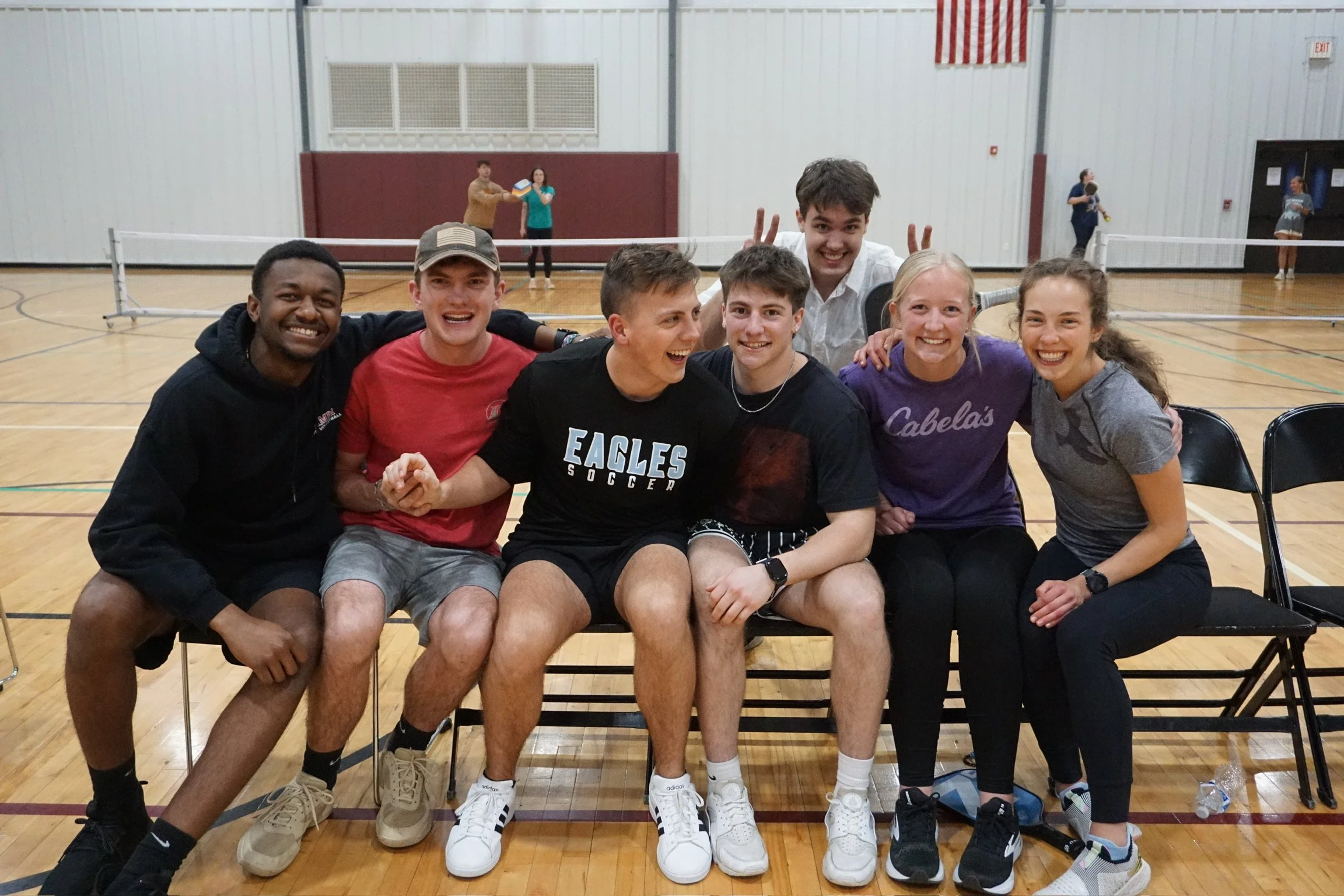 Group of seven young people sitting and standing in a gymnasium, smiling and posing for a photo, with a volleyball net in the background.