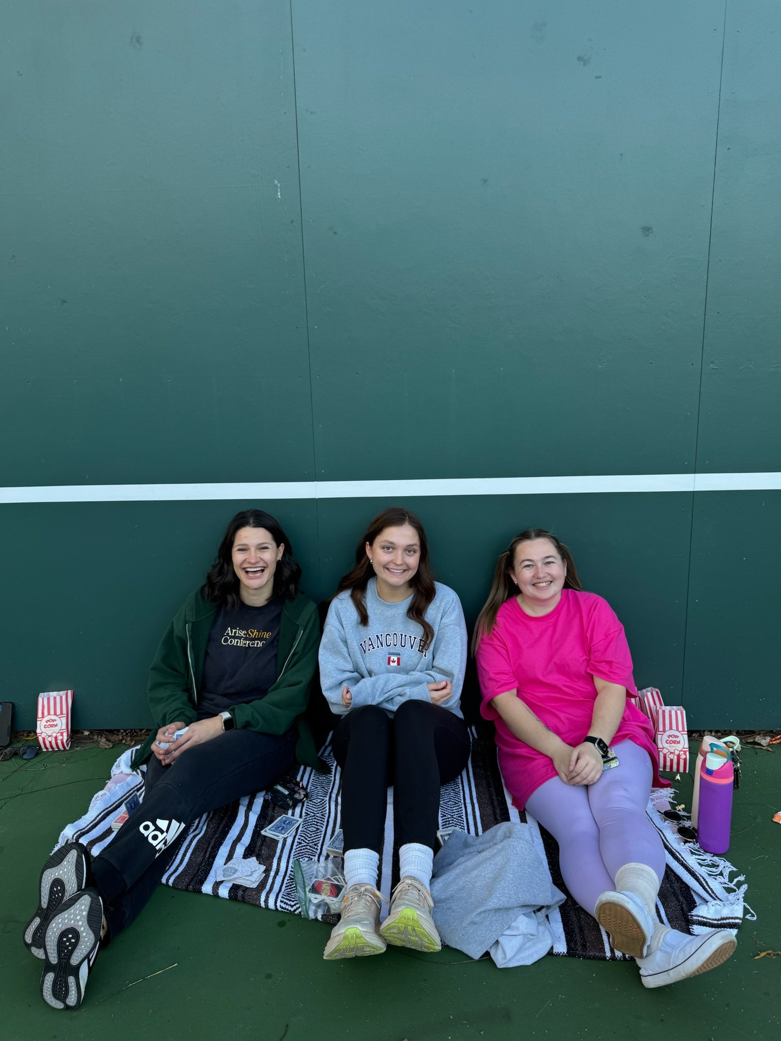Three young women sitting on a blanket against a green wall, smiling and enjoying each other's company.