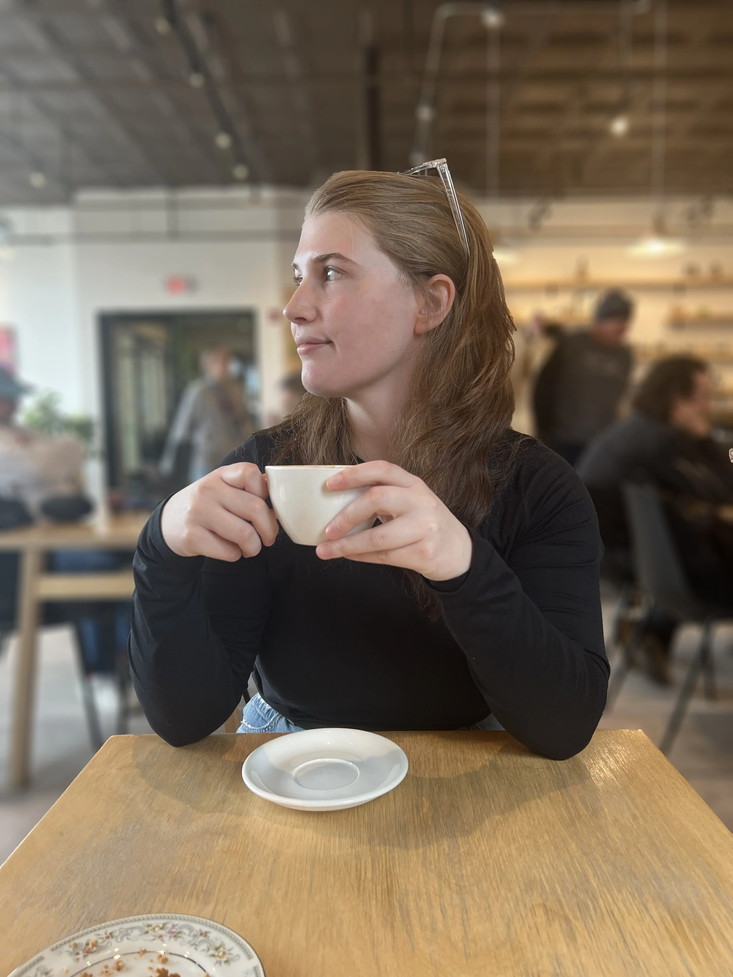 Woman with long brown hair and clear glasses resting on her head, holding a ceramic coffee cup with both hands, seated at a wooden table in a coffee shop.