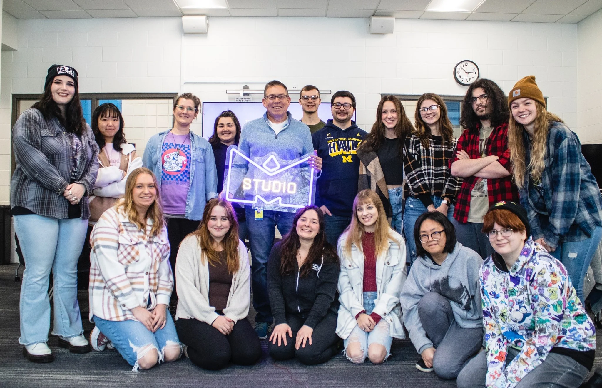 Group of diverse students and a teacher standing in a classroom, smiling for a group photo, with a neon 'Studio' sign and clock in the background.
