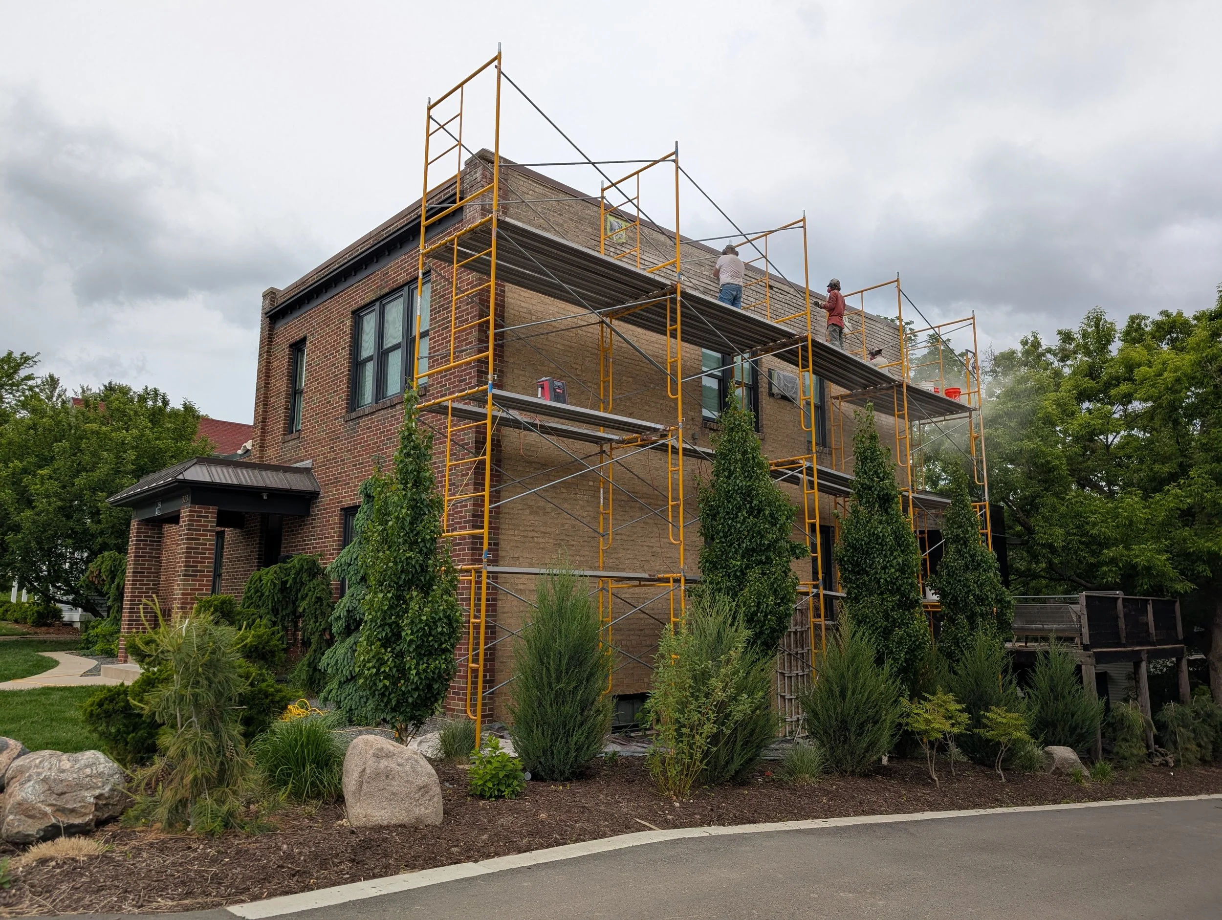 Two workers on yellow scaffolding working on the upper facade of a large brick house during daytime with cloudy sky. The house is surrounded by landscaped bushes and trees, and a curved driveway is in the foreground.