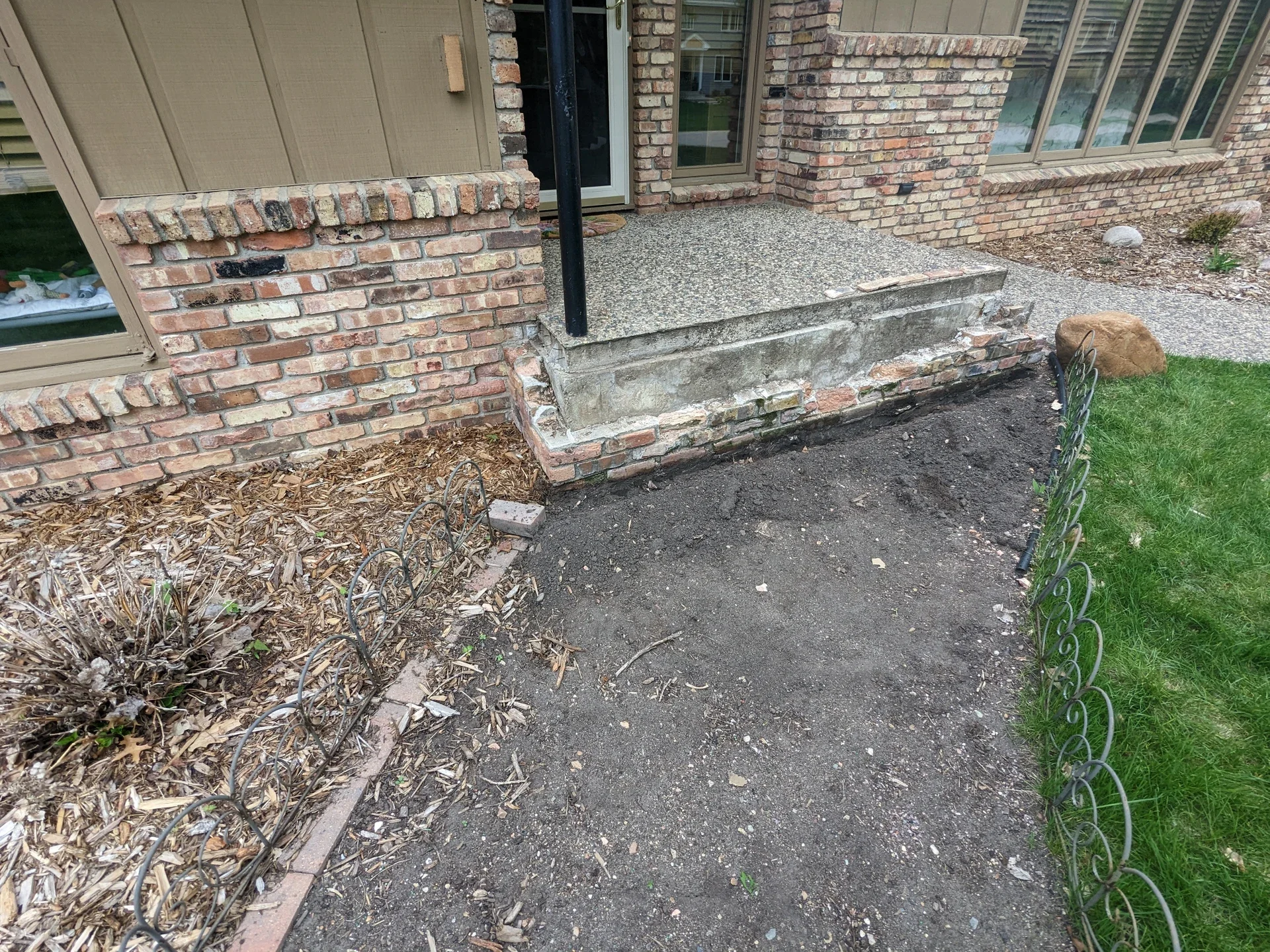 A small front yard area with a step leading up to a porch, with a black downspout. The yard is partially prepared with soil, surrounded by a decorative wire fence, and has some mulch and a large rock.
