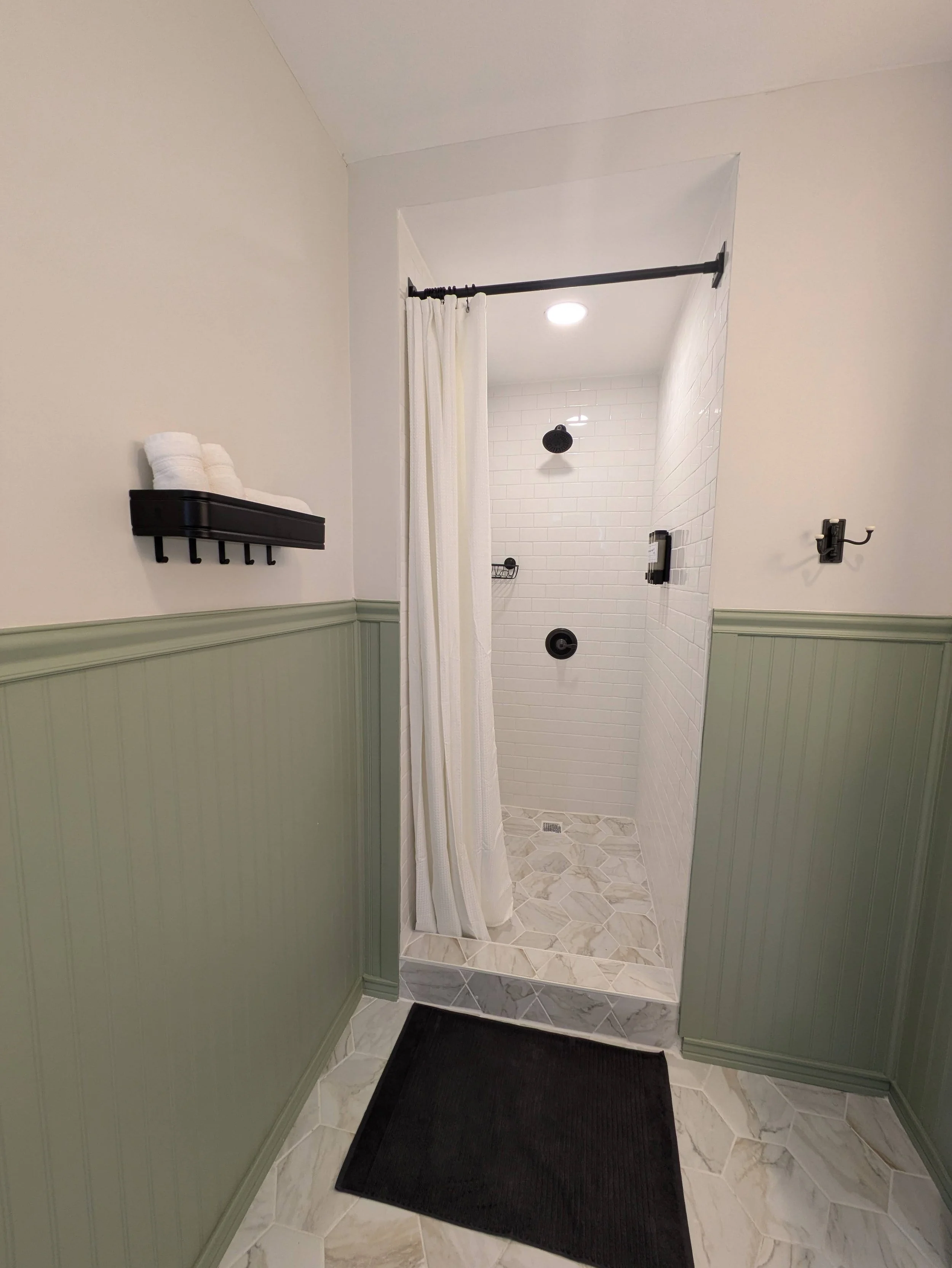 Bathroom with walk-in shower featuring white subway tile, black fixtures, and a beige and white patterned tile floor. A black shelf with folded white towels is on the wall, and a black hook is on the right wall. A black mat is on the floor outside the shower.