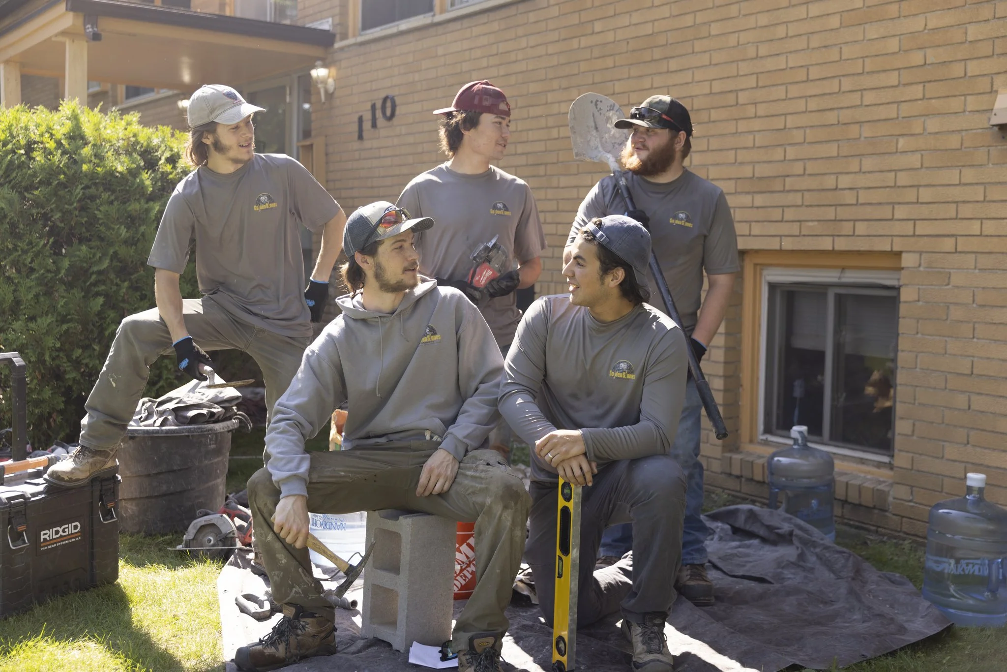 Six young men in work clothes gather outside near a brick house, engaging in conversation and taking a break from construction work with tools and building supplies around them.