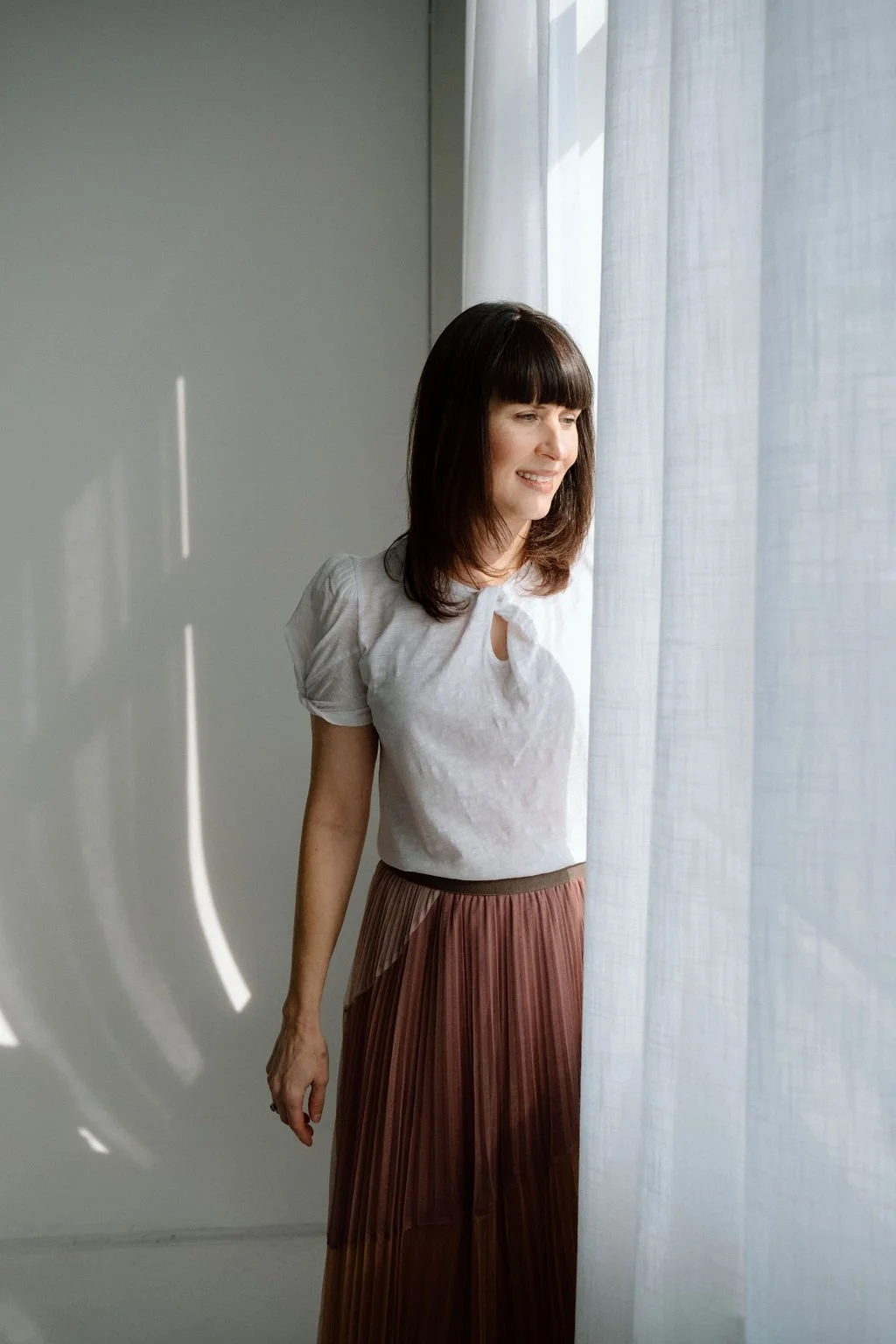 Woman with dark hair and bangs looking out a window with sheer curtains, smiling softly, wearing a white blouse and a brown pleated skirt.