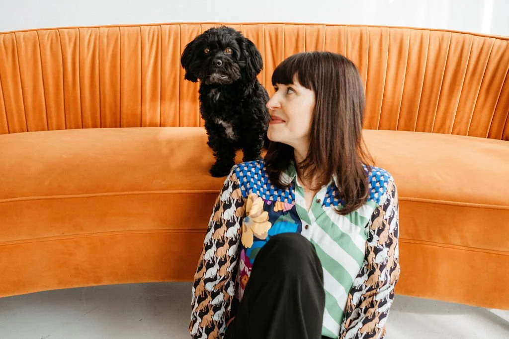 A woman with shoulder-length brown hair and a patterned shirt sitting on the floor beside a curved orange sofa. A small black dog is sitting on the sofa, looking at the camera.