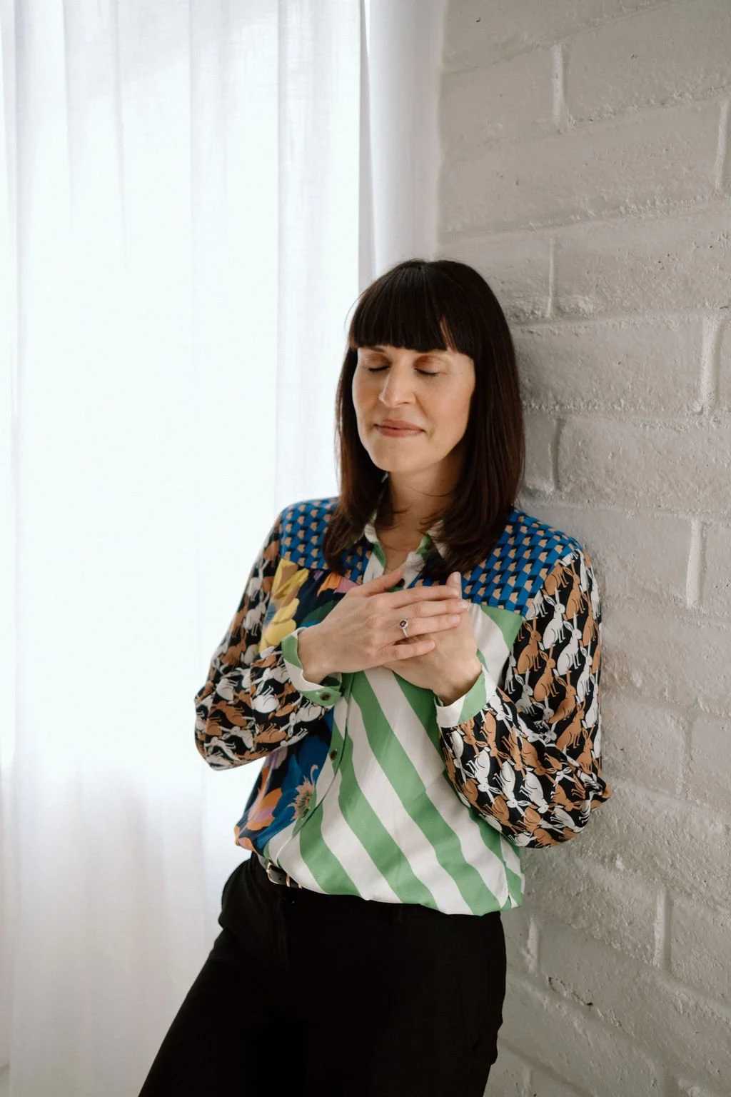 A woman with dark hair and bangs, wearing a colorful patterned shirt, stands with her eyes closed and hands on her chest, leaning against a white brick wall near a window with white curtains.