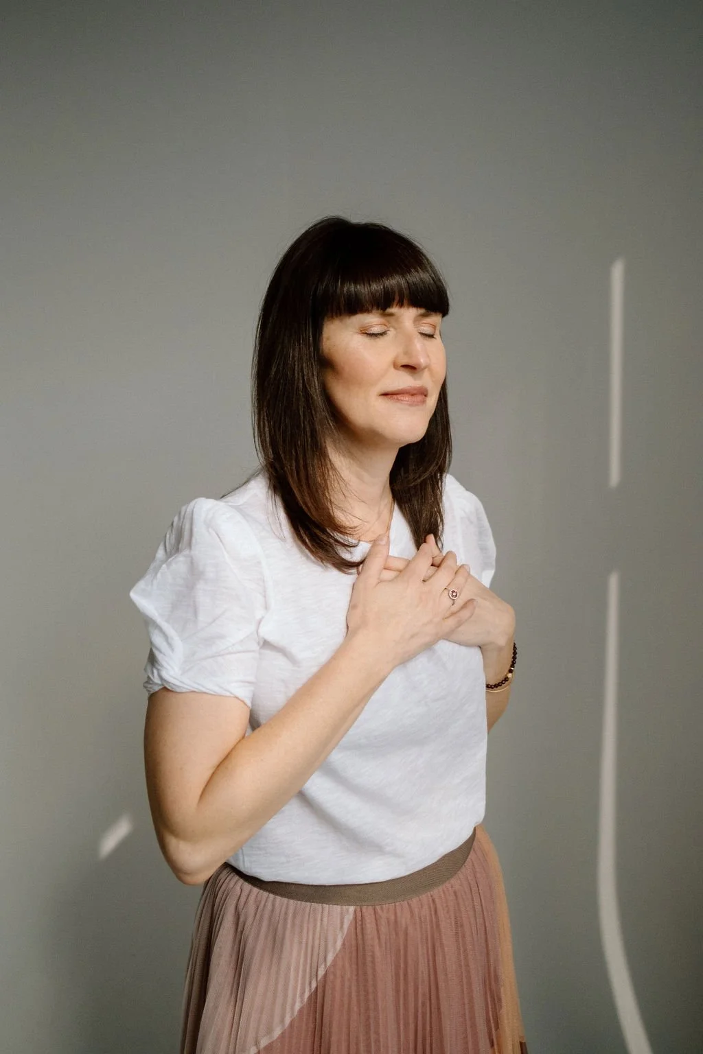 Woman with dark brown hair and bangs, wearing a white shirt and pink skirt, with eyes closed and hands on her chest, appearing to be praying or meditating.