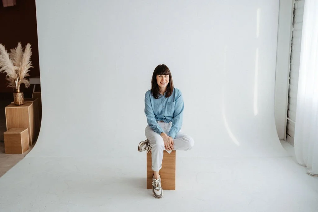 A woman with dark brown hair and bangs, smiling, wearing a light blue shirt, white pants, and sneakers, sitting on a wooden block in a photography studio with a white backdrop and natural light.