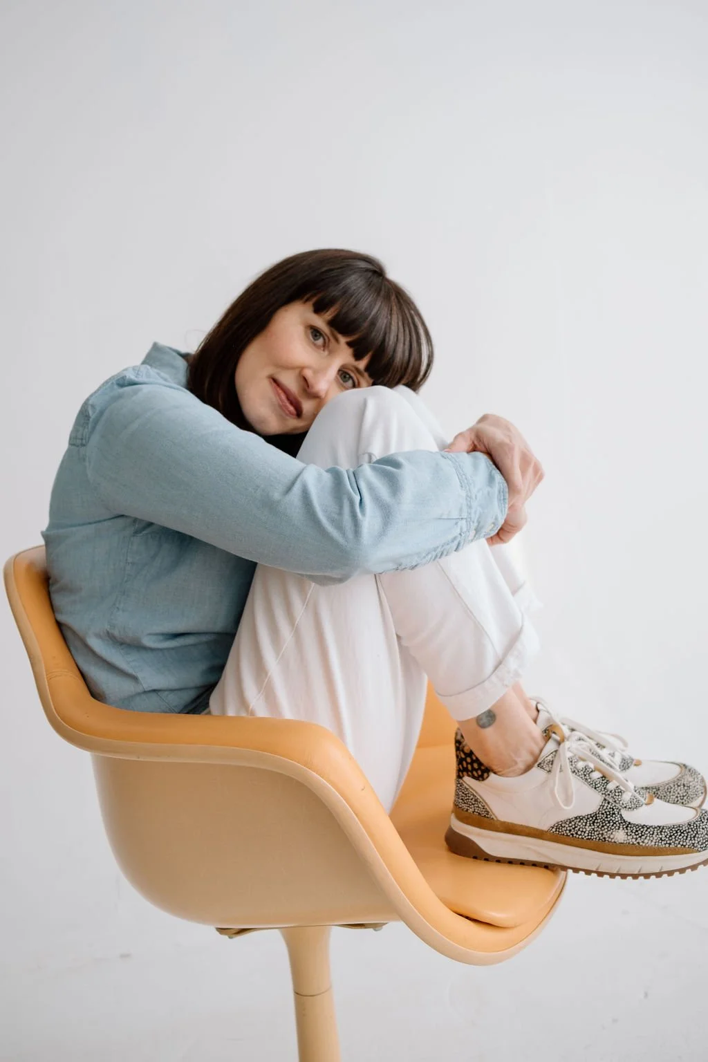 Woman sitting on a modern beige chair with her knees up to her chest, wearing a light blue shirt, white pants, and sneakers, smiling and leaning her face on her knees against a plain white background.