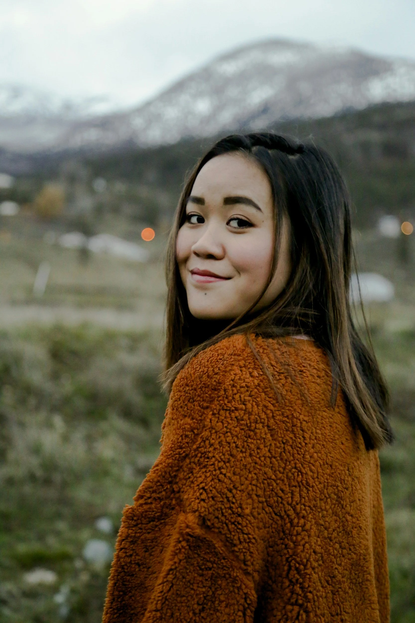 Young woman with long dark hair smiling outdoors wearing a rust-colored fuzzy sweater, with a mountain range in the background.