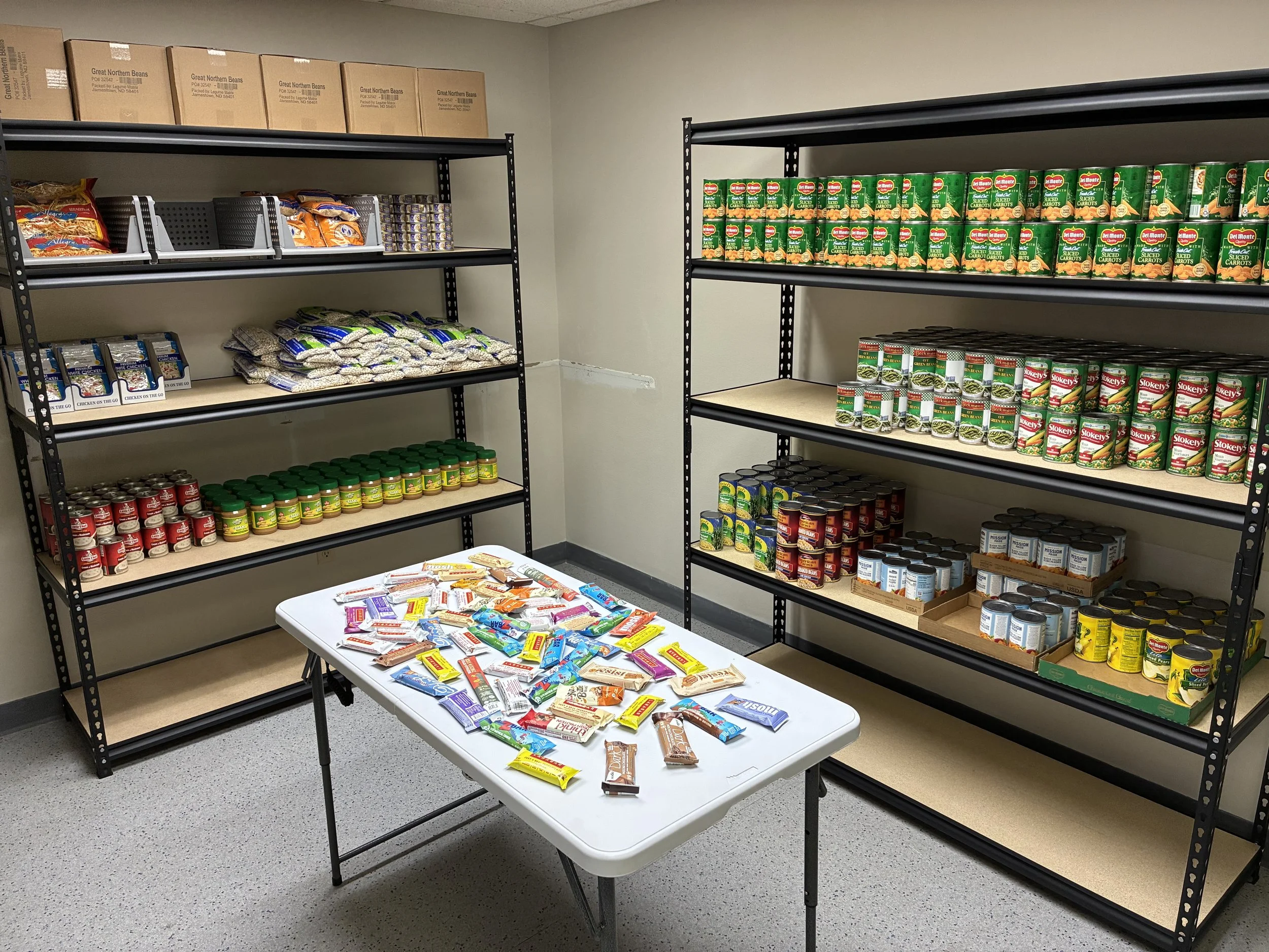 Shelves stocked with canned vegetables, beans, and other food items, with a table in front displaying packets of snack foods.