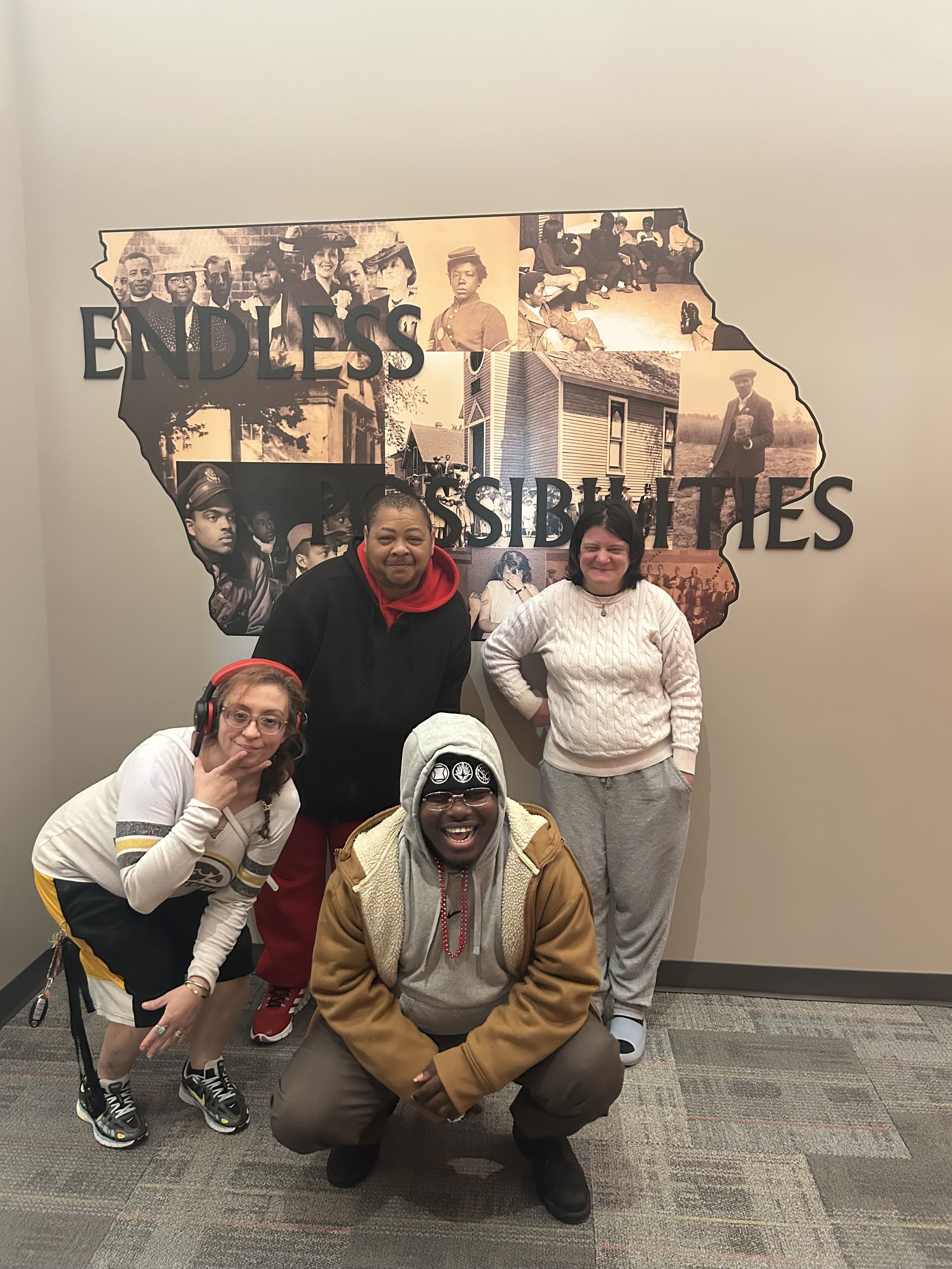 A group of five people posing in front of a wall with a mural of historical Black and white photographs of African Americans. The mural is shaped like the outline of the state of Georgia. The group includes two women and three men, all smiling and looking at the camera.