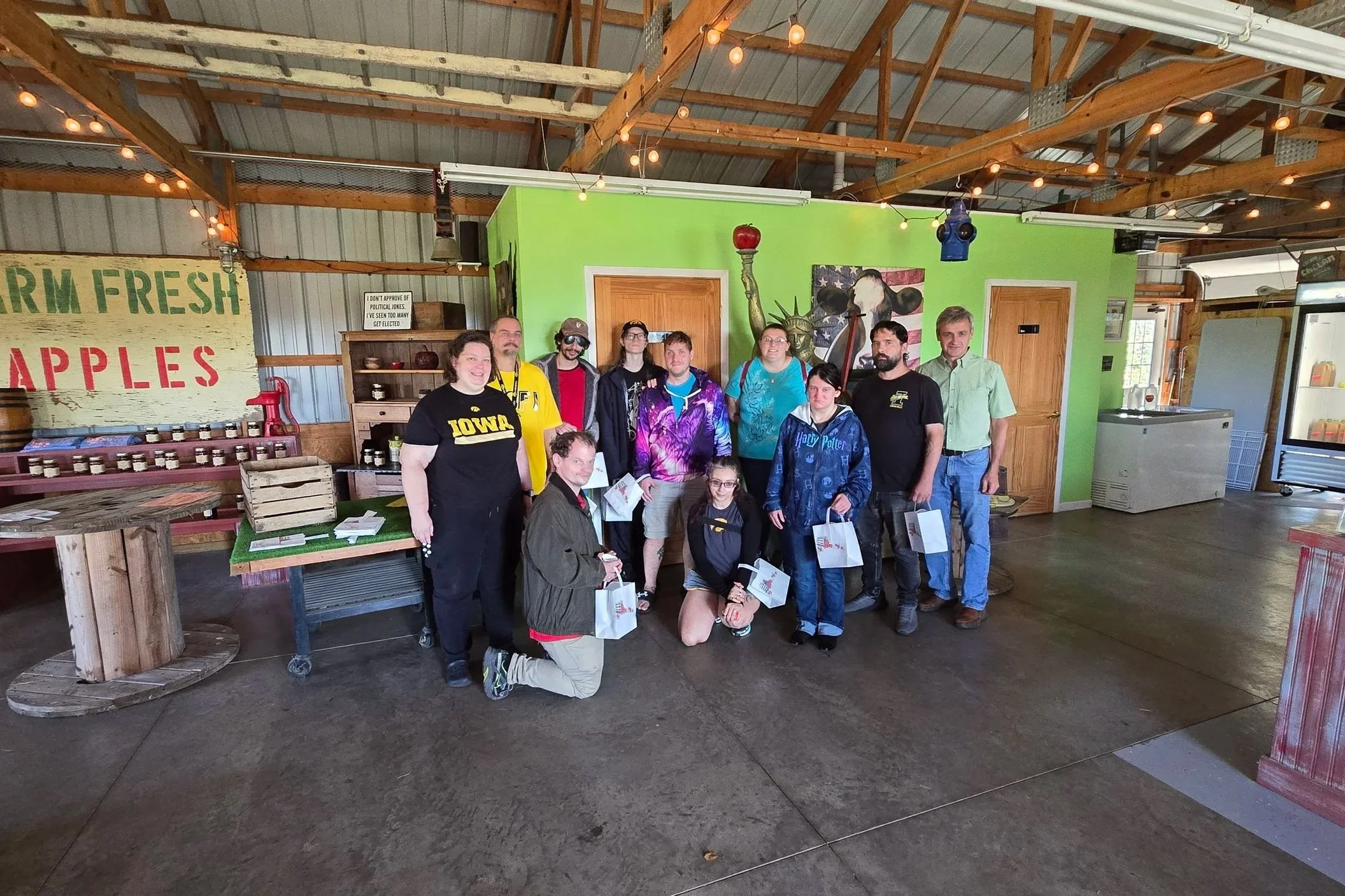 A group of twelve people standing inside a rustic farm-themed restaurant or market. The background features a green wall with farm art and shelves of jars, and a sign that says "Farm Fresh Apples." The group is holding shopping bags and some are smil