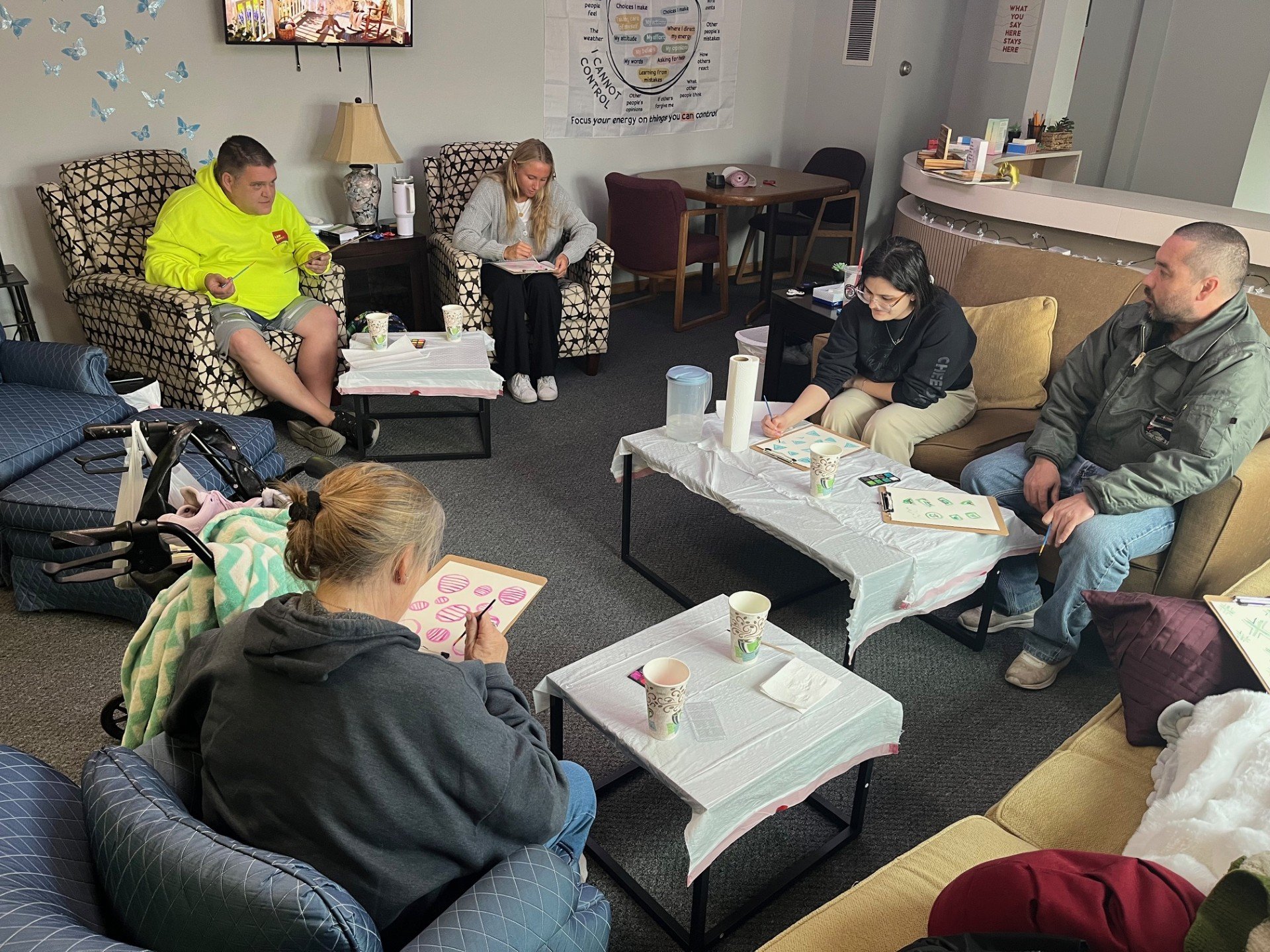 A group of people sitting in a room participating in a meeting or workshop. Four women and one man are visible, some are writing or drawing on paper. There are tables with cups, paper towels, and other materials. The room has a decorated wall with butterflies, a monitor, and a poster with phrases about control and attitude.