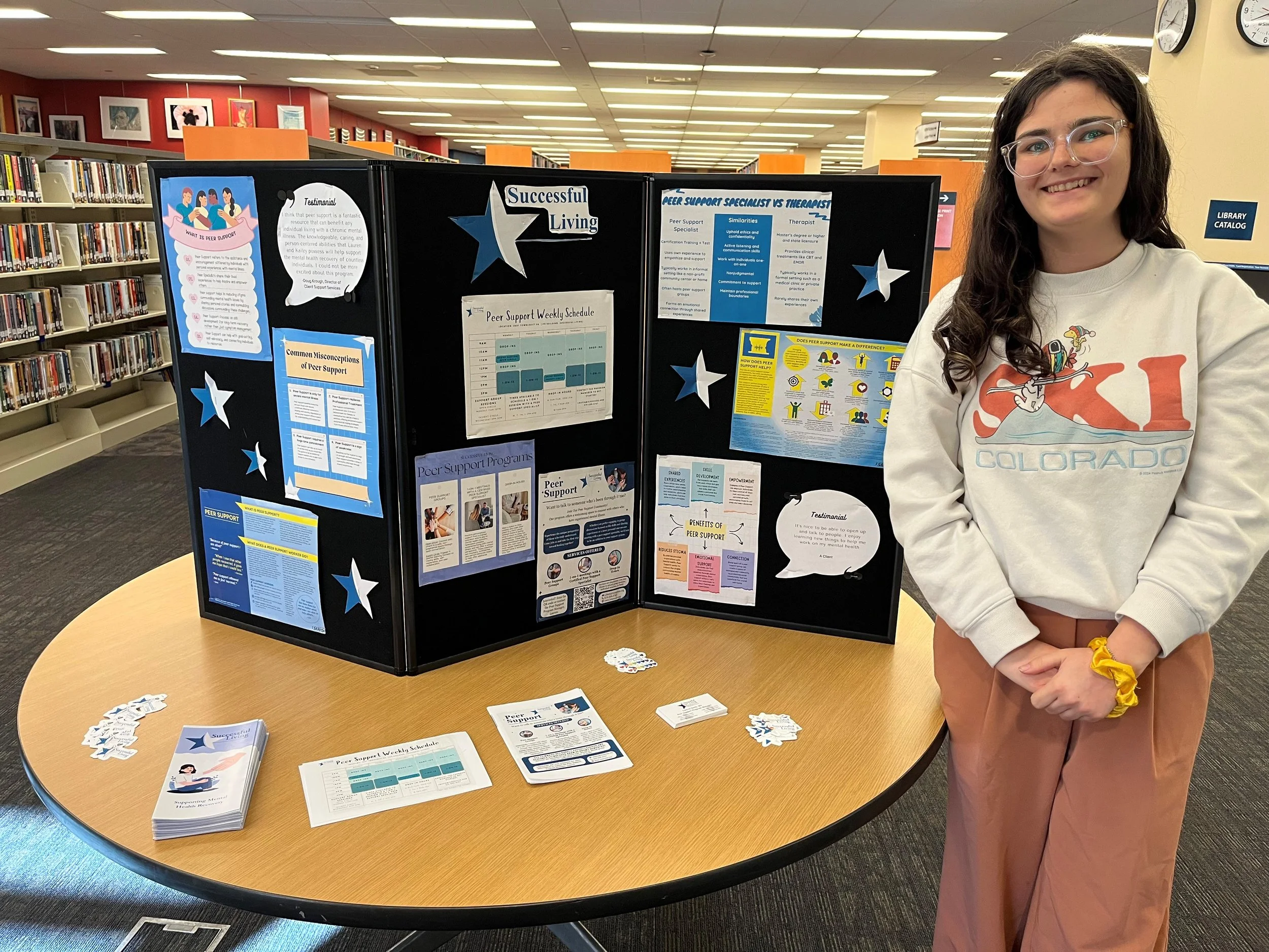 A young woman with long dark hair, glasses, and a light-colored sweatshirt stands next to a display board titled 'Successful Living' at a library. The display board contains informational posters and flyers on peer support programs. The library has bookshelves in the background and a clock on the wall.