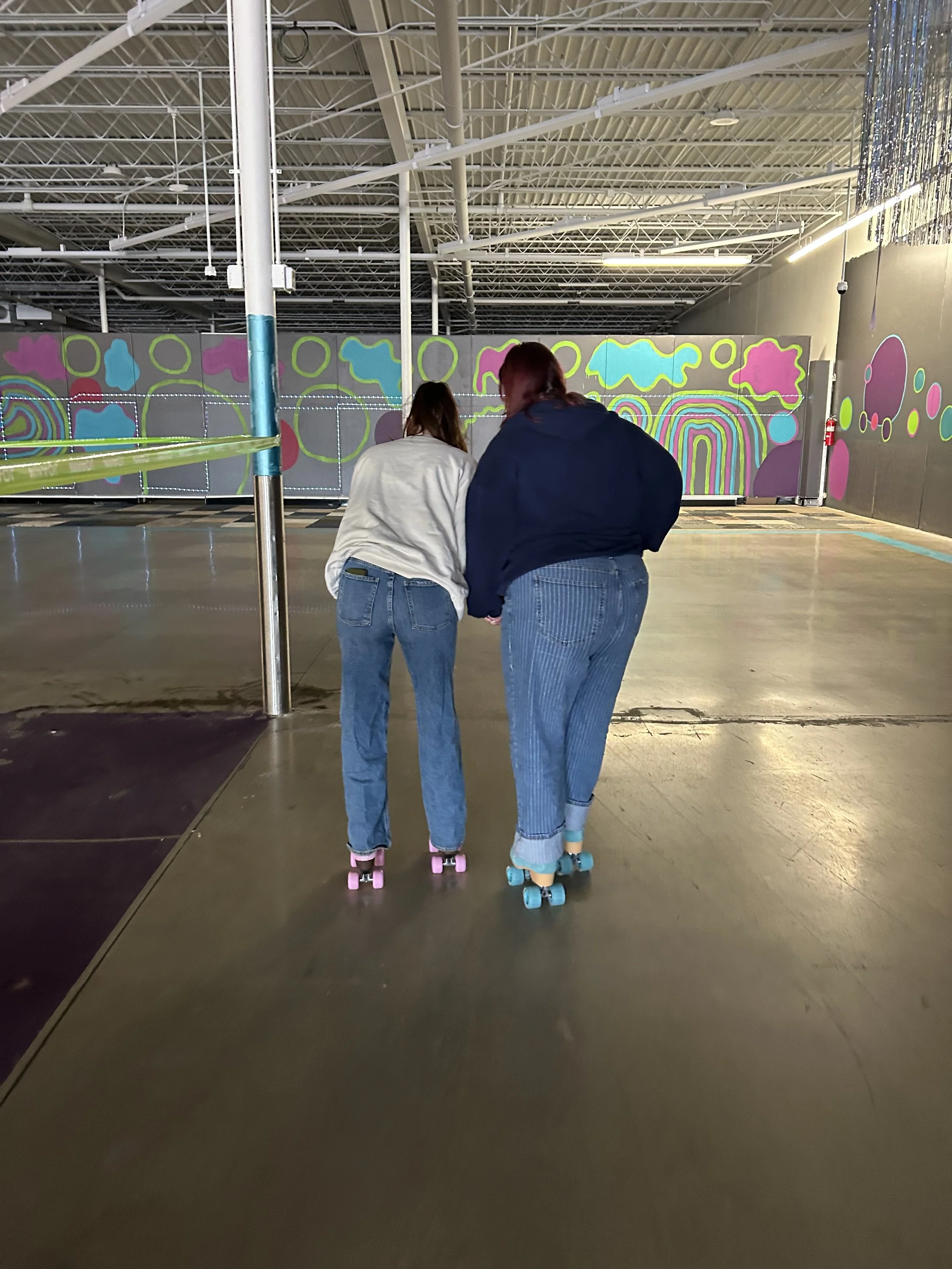 Two women roller skating in an indoor skating rink with colorful wall art in the background.