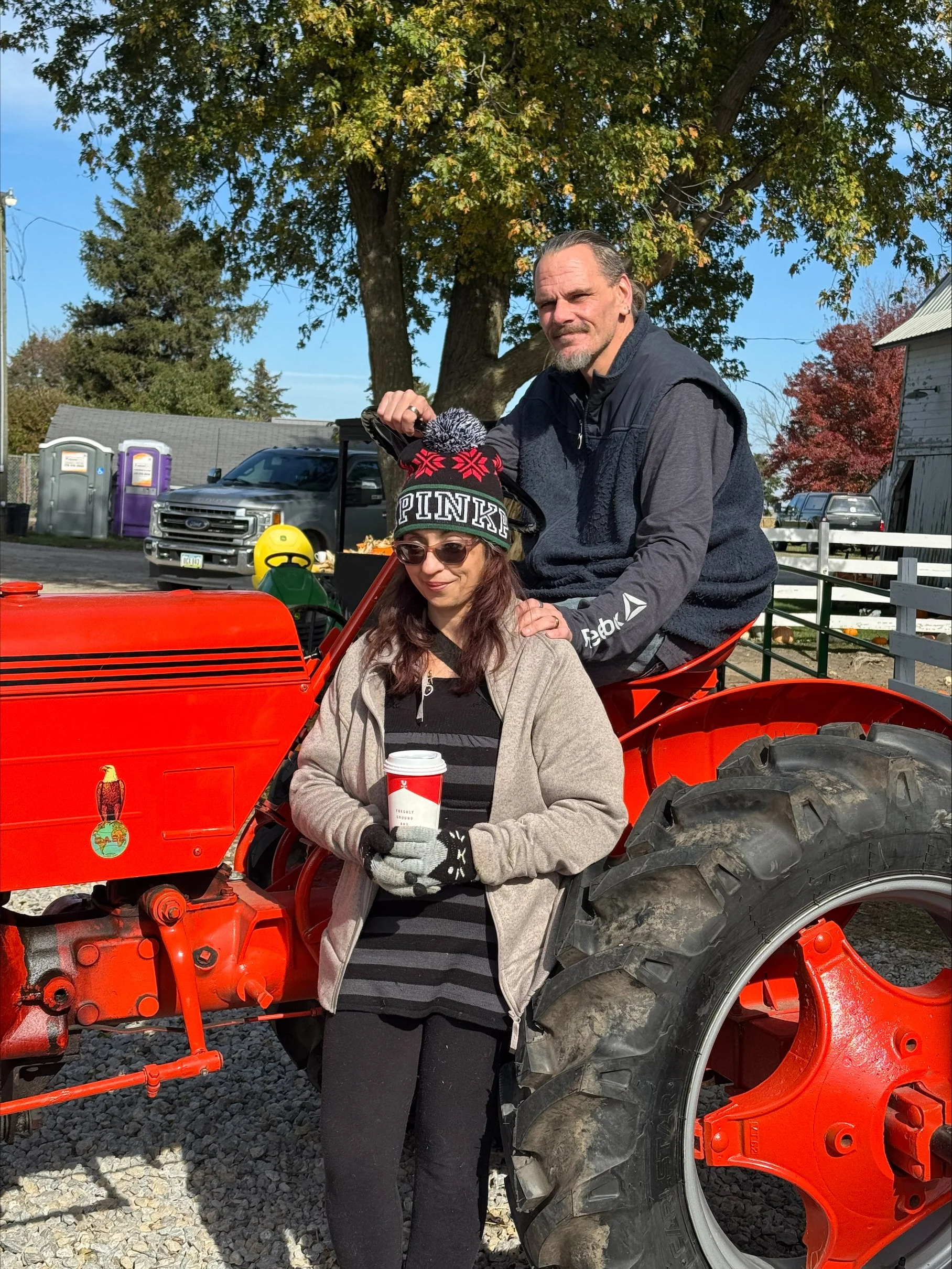 A man and a woman at an outdoor event, standing on a red tractor. The woman is holding a cup, wearing glasses, gloves, a beige jacket, and a striped shirt. The man is standing behind her, wearing a dark hoodie and vest, with his hand on her shoulder. There are trees and vehicles in the background on a sunny day.