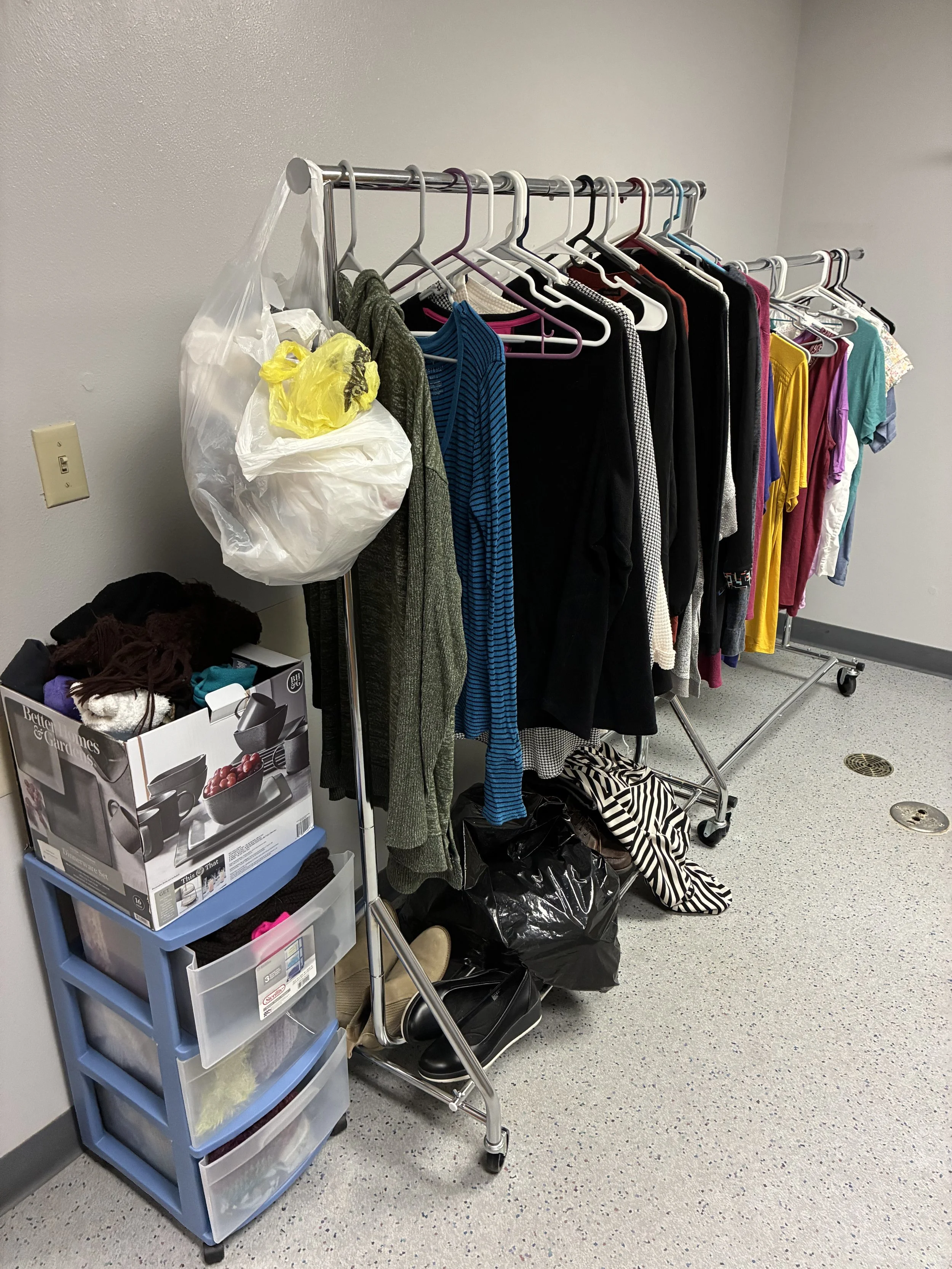 Clothes on a rolling rack, a plastic bag hanging from the rack, and a plastic storage drawer unit with folded clothes and a box of kitchen items, set against a plain white wall in a room with a light-colored speckled floor.