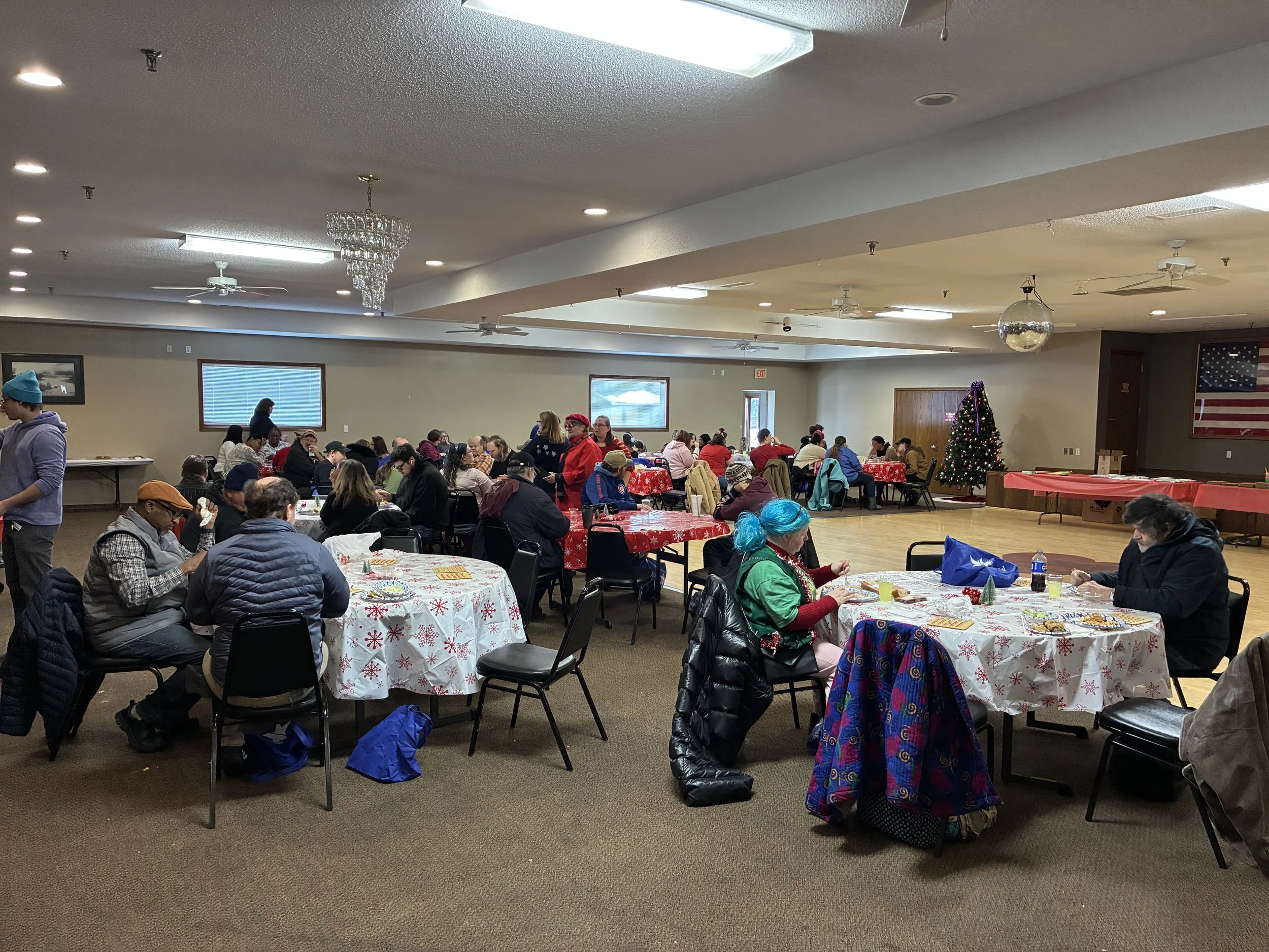 A large gathering of people inside a community hall decorated for the holidays with a Christmas tree and festive tablecloths. Attendees are seated at tables, some wearing holiday-themed clothing, with windows and American flag on the wall.