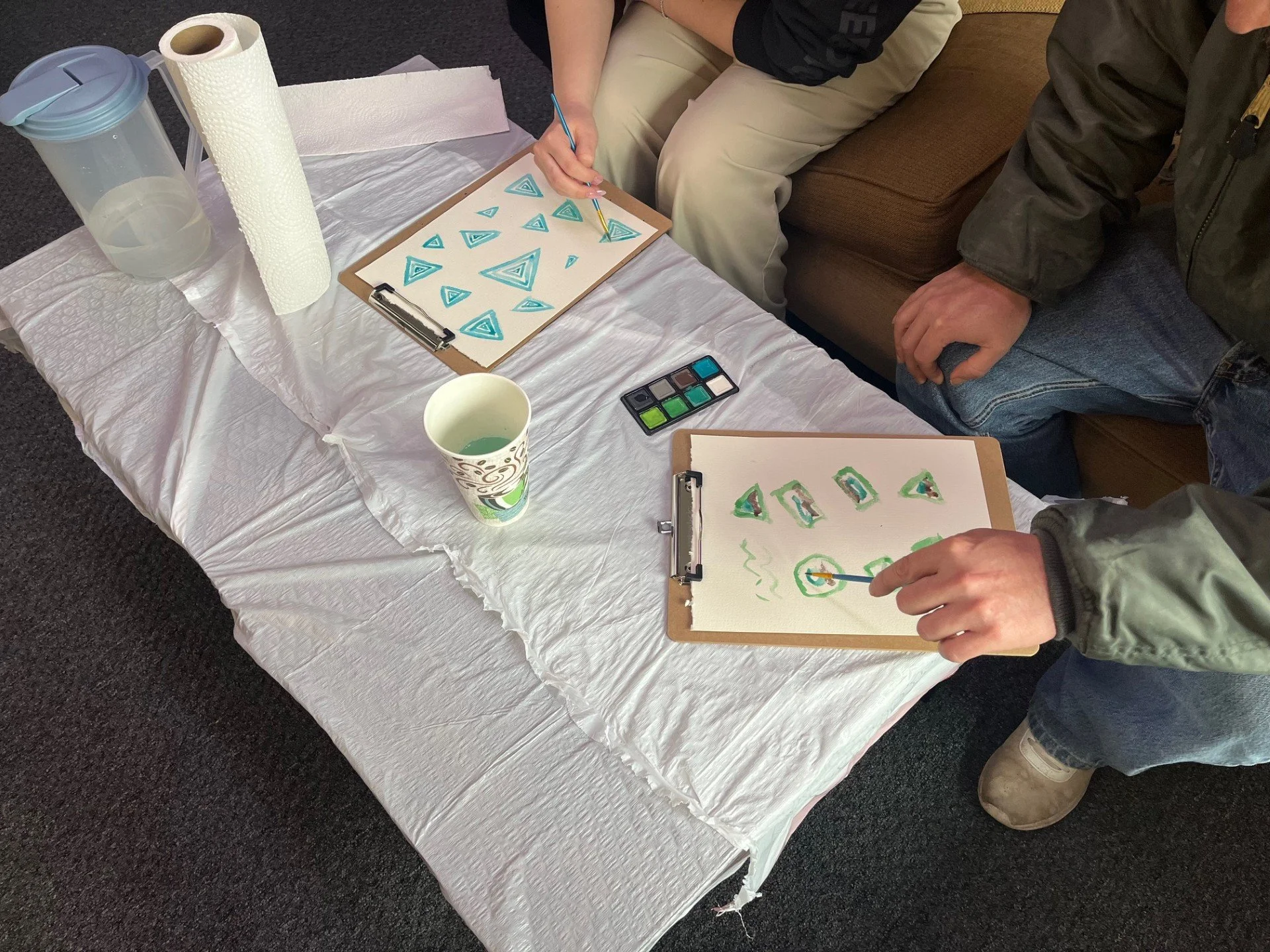 People creating art with watercolor paints on white paper, featuring geometric patterns and triangles, on a donation or charity event table covered with a white tablecloth, with a water pitcher, paper towels, and a paper cup nearby, in an indoor setting.