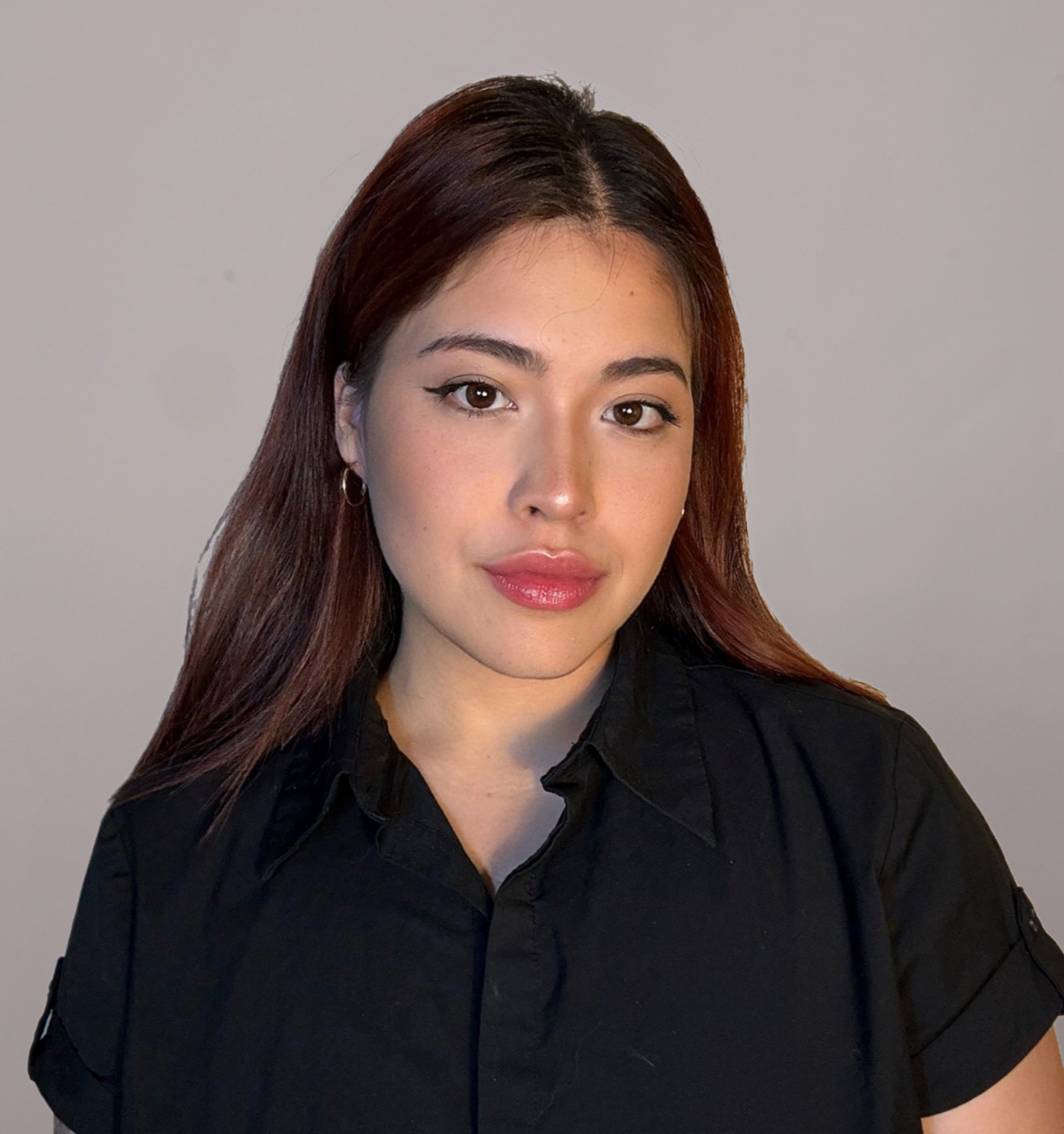 A young woman with long brown hair, wearing hoop earrings and a black shirt, looking into the camera against a plain gray background.