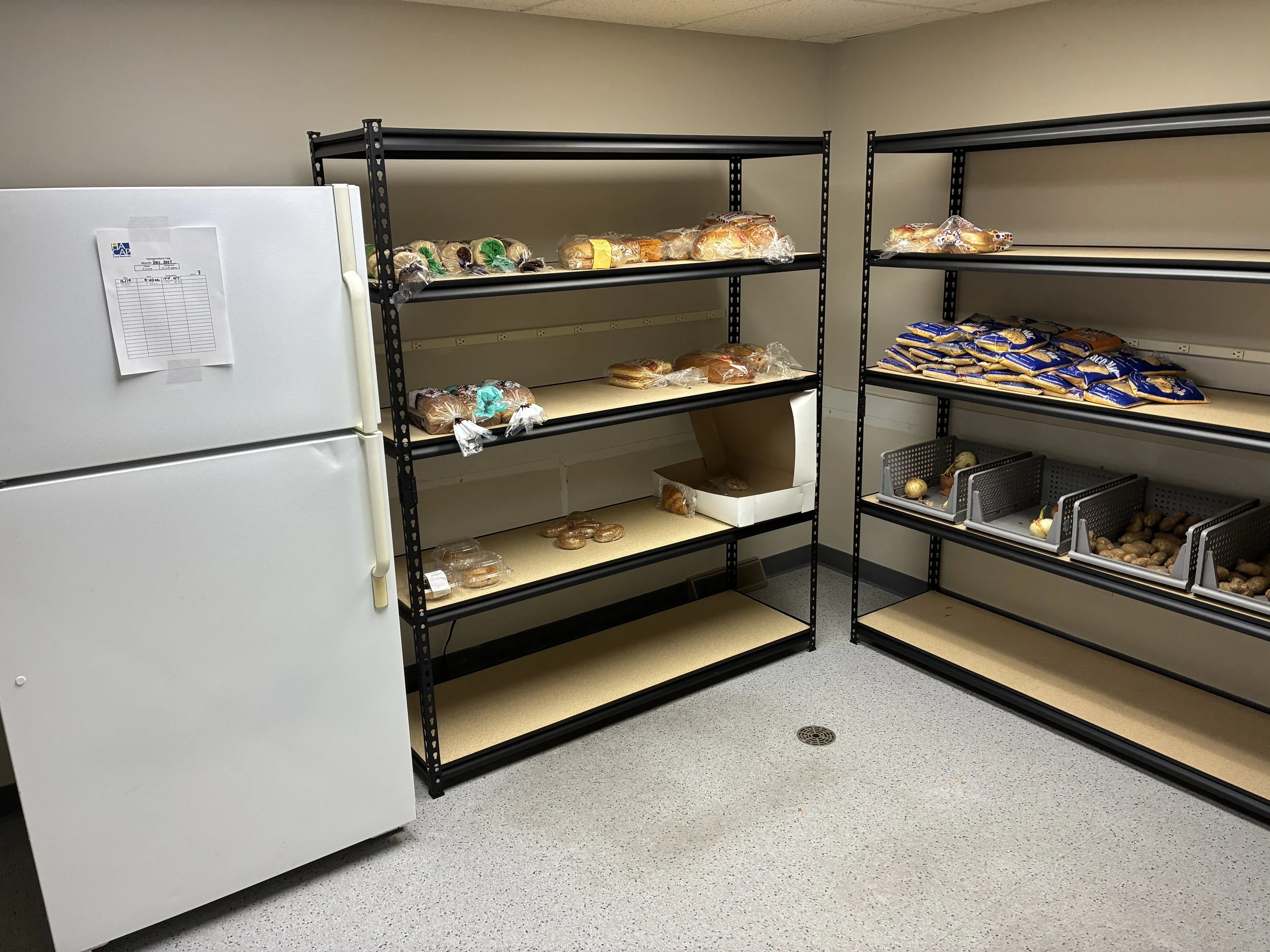 Empty metal shelving units in a room with a white refrigerator on the left, some packaged baked goods and potatoes on the shelves.