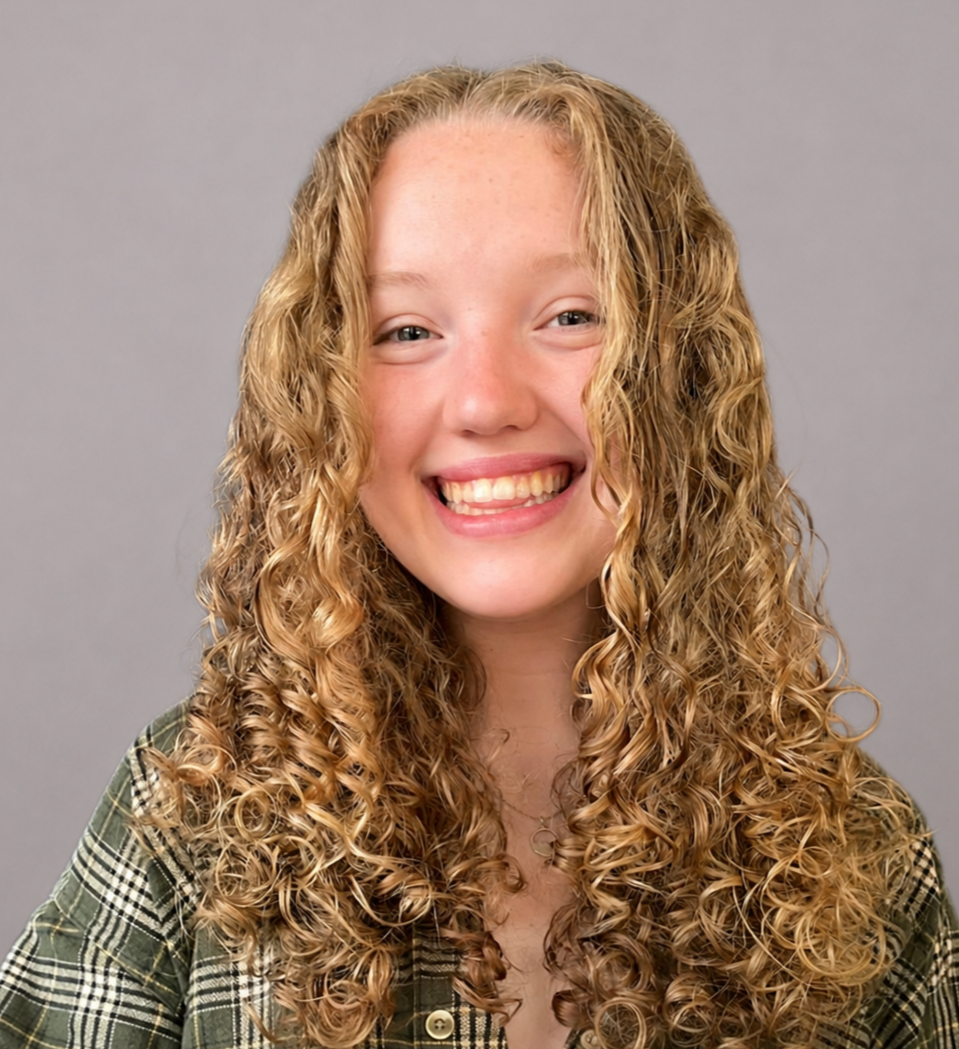 A young woman with long, curly blonde hair smiling against a gray background.
