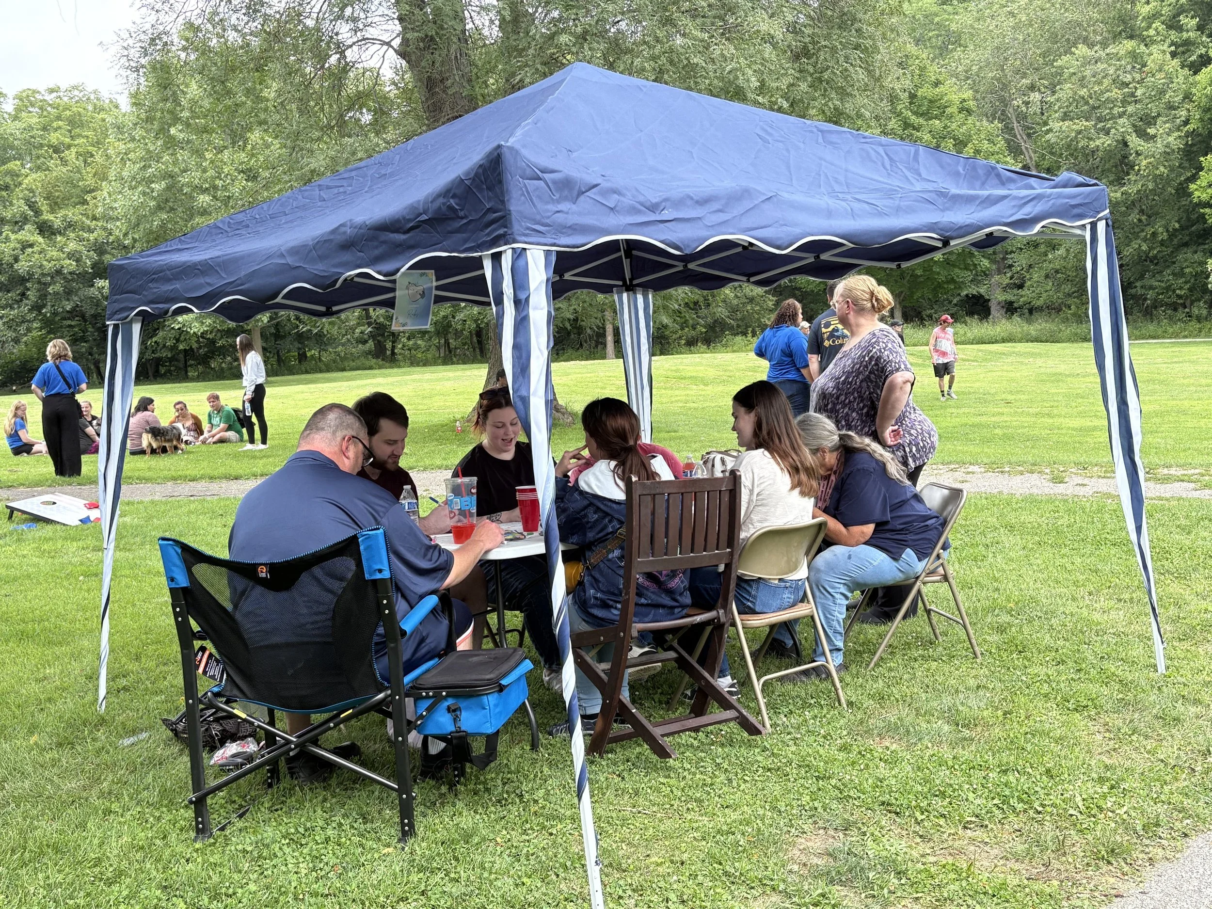 A gathering of people sitting and standing around a table under a blue canopy in a grassy park, with many trees in the background. Some people are talking, and a few are looking at their phones.