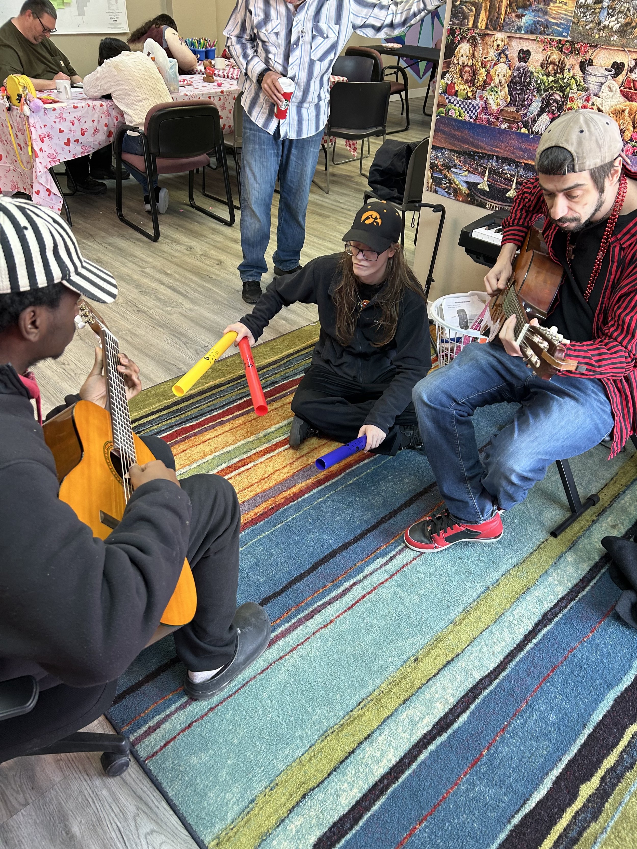 Group of people playing guitars and holding colorful plastic tubes in a room with a colorful striped rug and wall art.