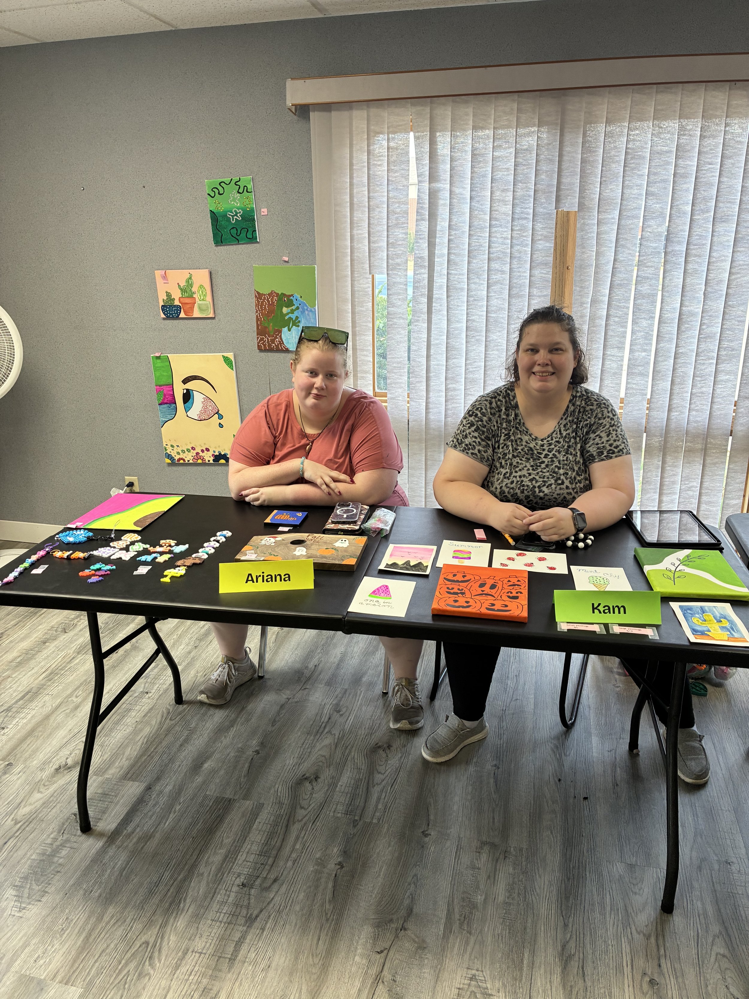 Two women sitting at a table with various artwork and crafts displayed. One woman has glasses on her head, wearing a pink shirt, and the other has short curly hair, wearing a leopard print shirt. They are in a room with gray walls and vertical blinds