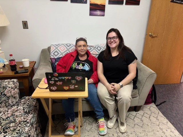 Two women sitting on a beige couch in a room, smiling at the camera. The woman on the left has gray hair, red jacket, and rainbow sneakers, with a laptop decorated with hearts on her lap. The woman on the right has long dark hair, glasses, black shirt, and white pants. There is a small wooden table with drinks and snacks beside them and pictures hanging on the wall above.