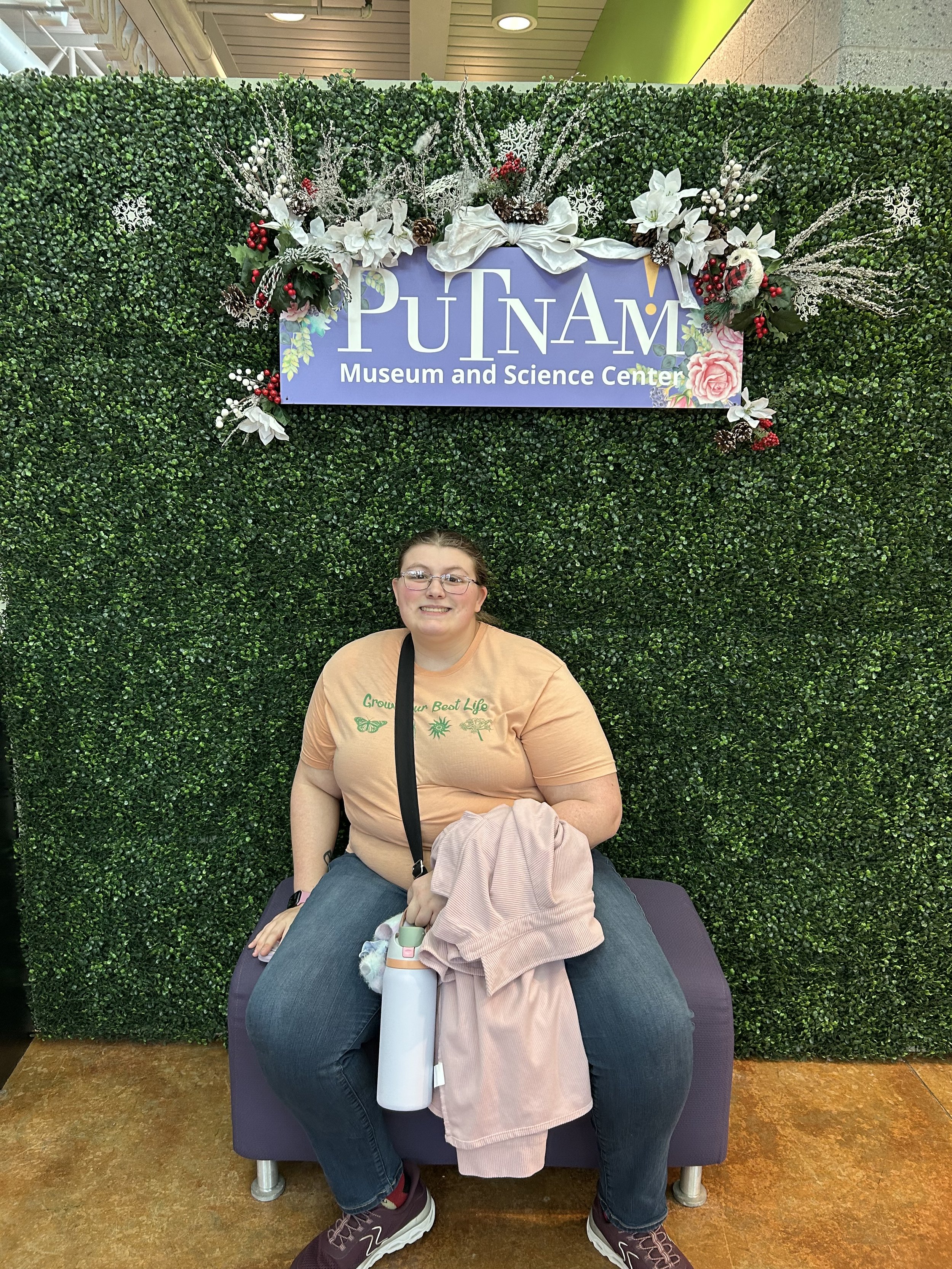 A woman with glasses and a beige T-shirt sitting on a purple chair in front of a green foliage wall with a sign that reads 'Putnam Museum and Science Center.' She is smiling, holding a light pink jacket, with a white water bottle on her lap.