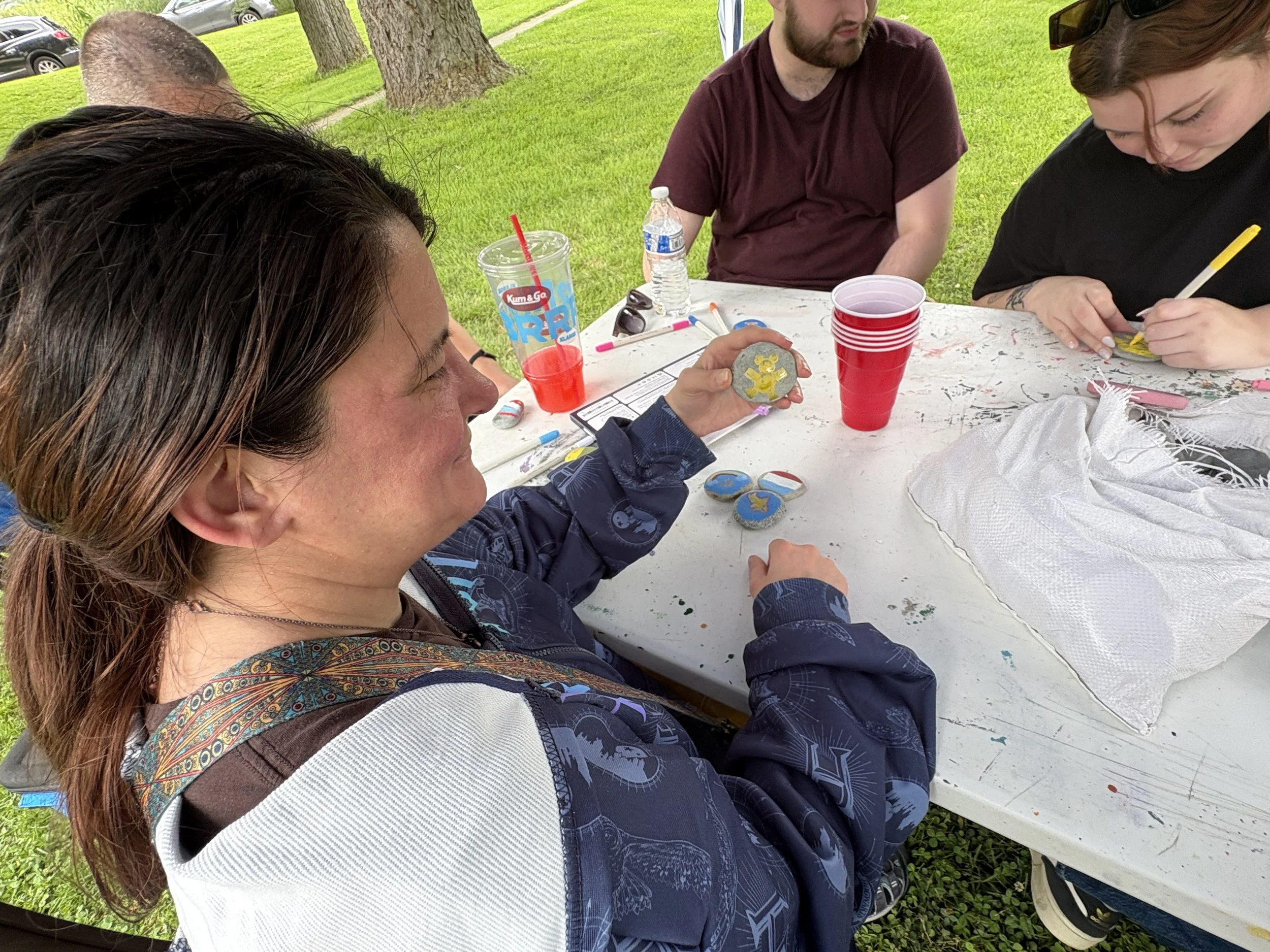 People gathered around a table outdoors painting rocks with colorful designs and messages, with grassy area and trees in the background.