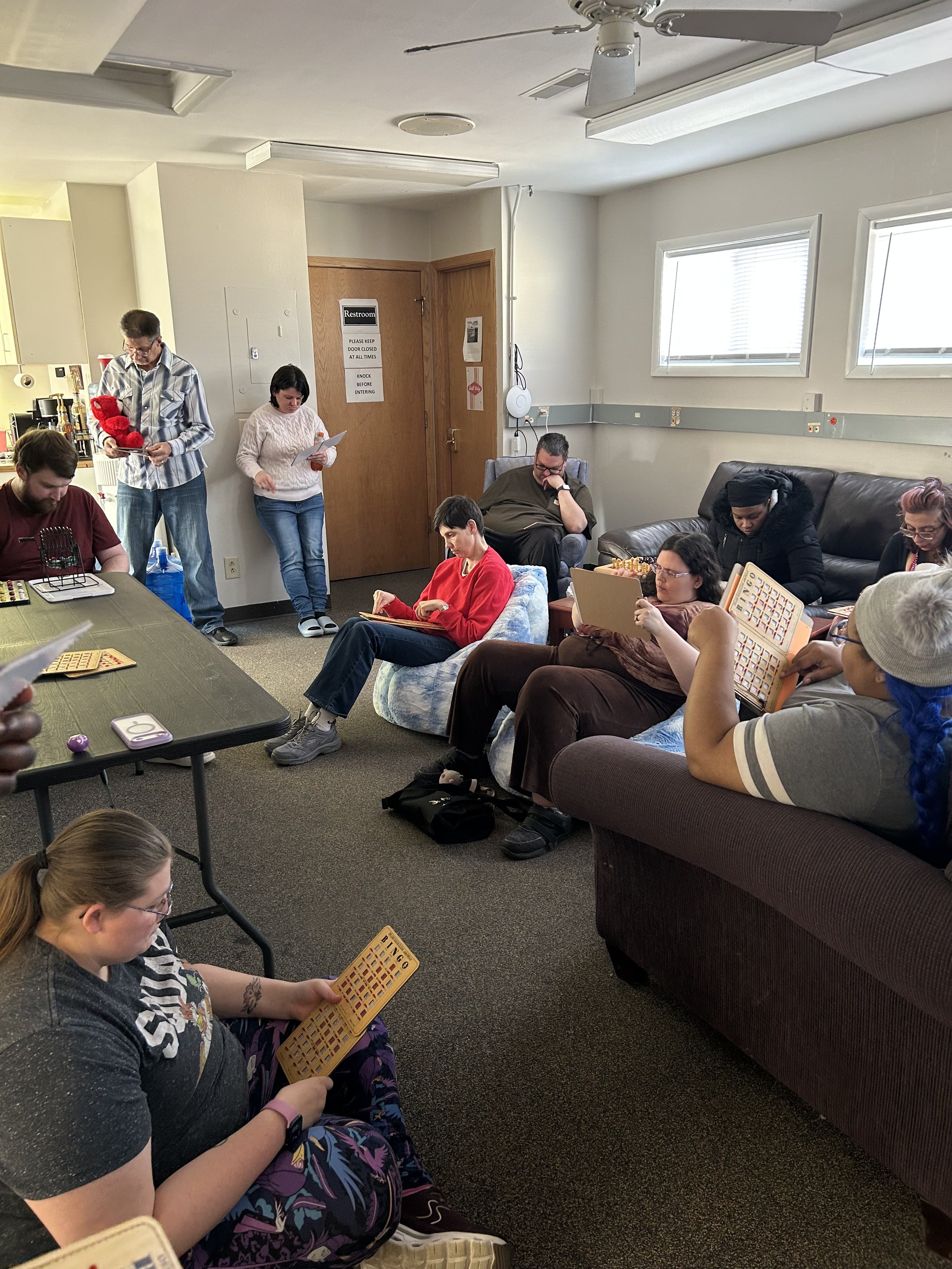 A group of people gathered in a room, sitting on couches and chairs, playing bingo with bingo cards. Some are reading their cards, while others look at papers or electronic devices. The room has windows, a ceiling fan, and a door with a sign that says Restroom.